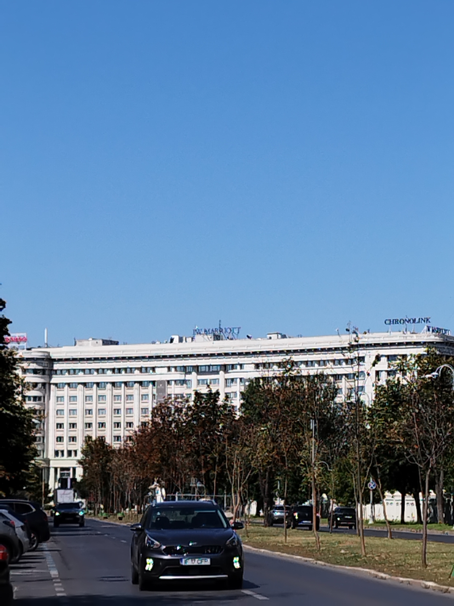 The image shows a street scene in Bucharest, Romania, featuring a large, white, multi-story building that appears to be a hotel. The building has a consistent facade with rows of windows. The name "JW MARRIOTT" is visible on the top of the building, indicating its identity. Another sign, "CHRONOLINK," is also present. The sky is clear and blue. In the foreground, there are several cars on the road, including a dark-colored SUV with a Romanian license plate "IF 17 CFP" in the right lane. Trees line the street, with some showing autumn colors. The overall scene depicts an urban environment on a clear day.