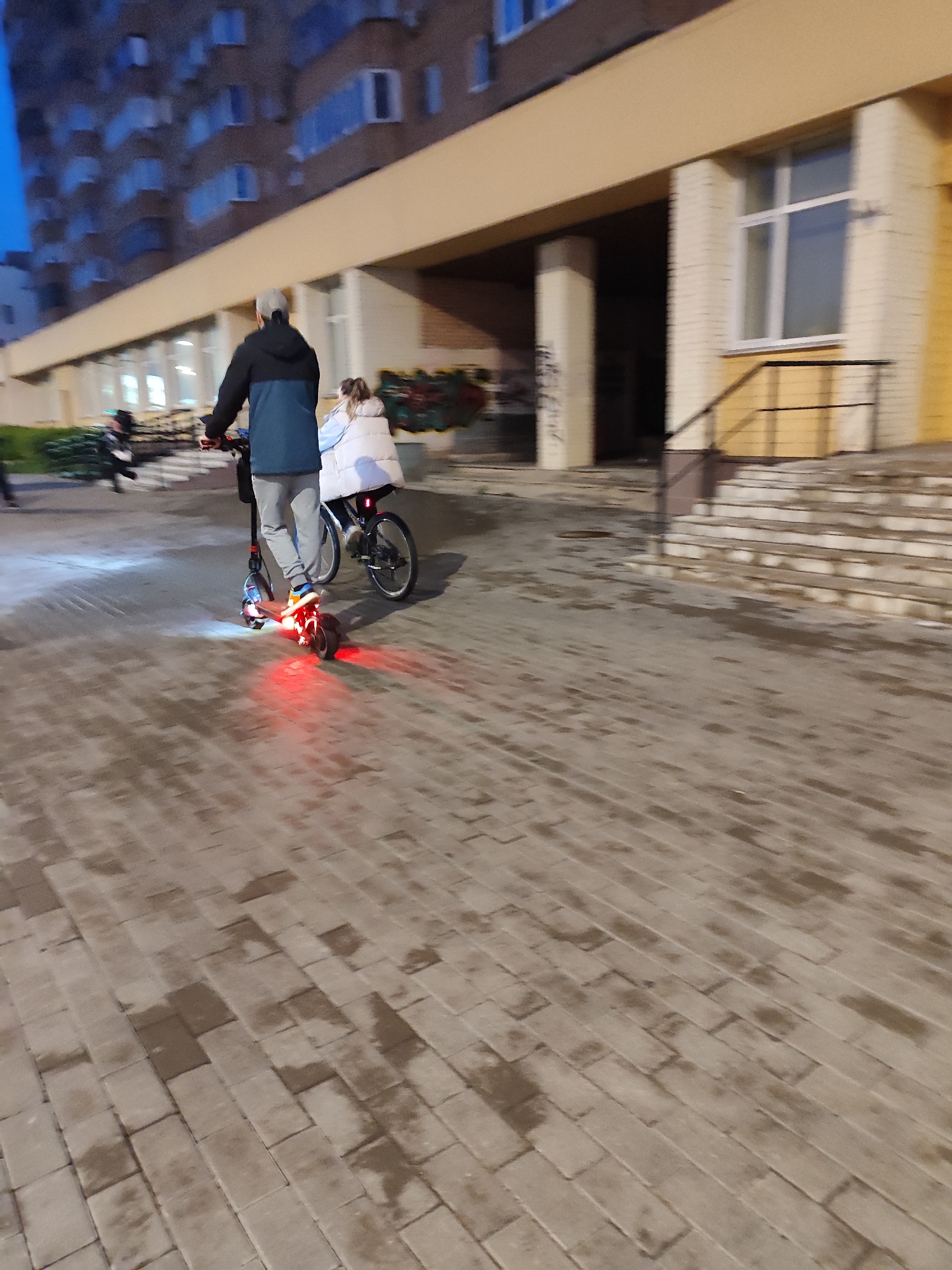 In an urban setting at dusk, two individuals are visible on a paved area in front of a building. The primary focus is on a person riding an electric scooter, who is moving away from the camera. This individual is wearing a dark jacket with a blue section on the sleeves and a light-colored cap. They are dressed in light gray pants and dark shoes with orange accents. The scooter has red lights illuminated on its rear.
Following closely behind the scooter rider is another person, seated on a bicycle. This individual is wearing a light-colored puffy jacket and appears to be either a child or a smaller adult. The bicycle has a dark frame and visible wheels.
The environment consists of a building with a light beige facade and a dark canopy. There are windows visible on the building, some of which are lit from within. A set of stairs with a railing leads up to an entrance. The ground is paved with rectangular stones that appear wet, reflecting some of the ambient light. The overall lighting suggests it is either early evening or late twilight. The background shows a tall residential building with multiple floors and windows. The scene is outdoors, on a sidewalk or pedestrian area.