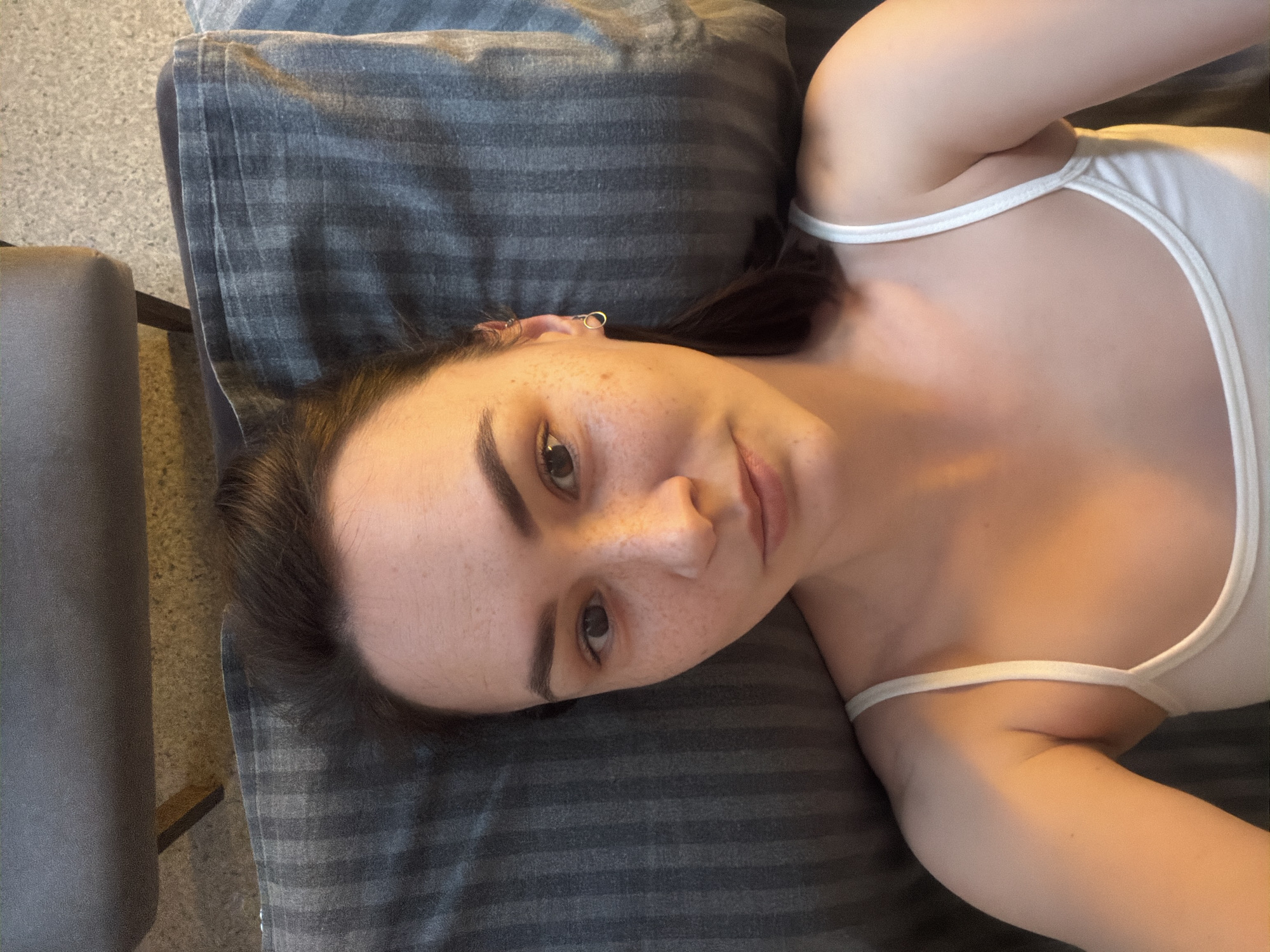 The image captures a young person, likely a woman, lying down and looking directly at the viewer. She is positioned diagonally on a dark grey pillow with subtle horizontal stripes. Her dark hair frames her face, and she has distinct dark eyebrows and numerous freckles scattered across her nose and cheeks. She is wearing a simple white camisole or tank top with thin shoulder straps. A small silver hoop earring is visible in her left ear. Her expression appears calm and neutral, conveying a relaxed mood. In the background, to the left, parts of a dark grey upholstered chair or furniture piece and a light grey, textured floor are visible. The lighting is soft and even, indicating an indoor setting. No specific time of day or weather conditions can be determined from the image, and no text is visible. The provided location context is Chaikovsky, Russia.