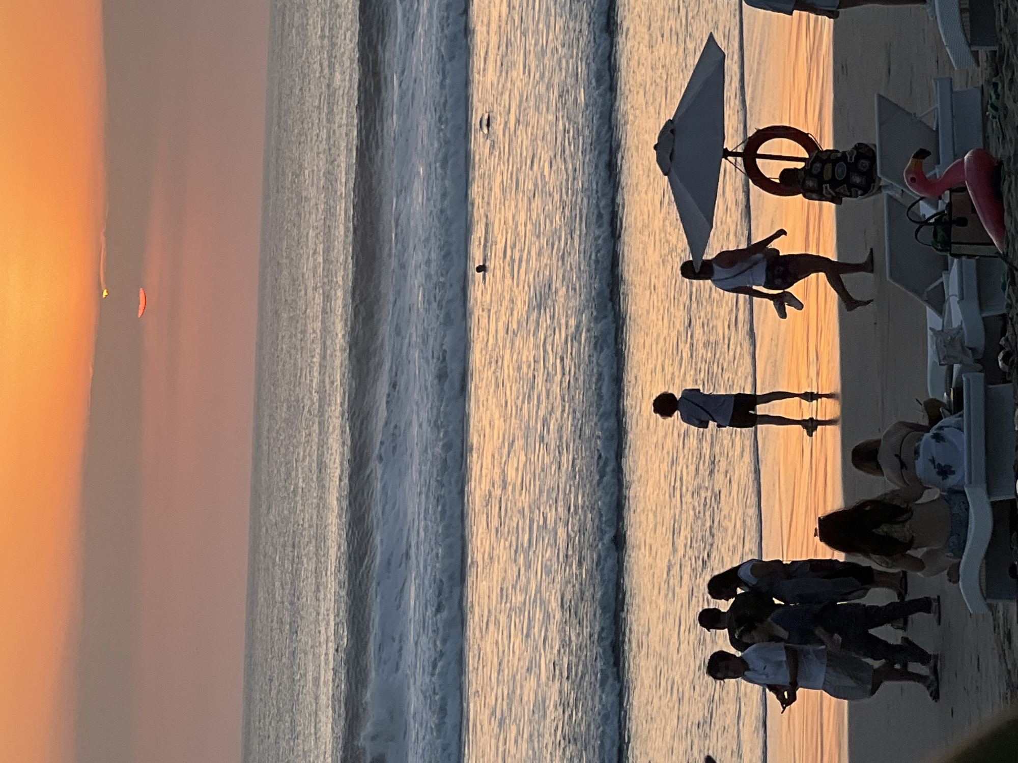 The image captures a sunset scene on the beach in Kuta, Indonesia. The sky is a vibrant orange, with the sun partially obscured by clouds, casting a warm glow on the ocean and wet sand. Gentle waves roll towards the shore, reflecting the sunset colors. Several people are visible on the beach and in the water. On the sand, a group of individuals are walking and standing, some appearing to be tourists. Further back, a woman walks under a beach umbrella, carrying flip-flops. Near the umbrella, a person is kneeling, and a life preserver is hanging. In the foreground, lounge chairs are set up, with a pink flamingo inflatable visible. Two individuals are seated on a lounge chair in the mid-foreground, facing away from the camera. In the water, a person is swimming.