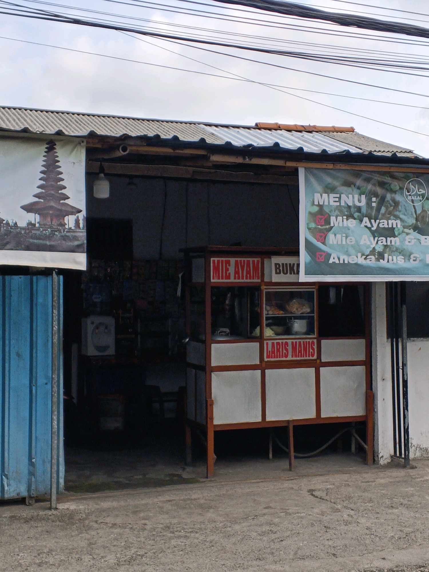 The image shows a street-level view of a small food stall in Gianyar, Indonesia. The stall has a simple, open-fronted structure with a corrugated metal roof. A banner above the stall displays "MENU : Mie Ayam" and "Aneka Jus & B". Another banner on the left side of the image features a picture of the Pura Besakih temple, a significant landmark in Bali, suggesting a connection to tourism or local culture.
In the foreground, a blue metal gate or door is visible on the left. The stall itself is a cart-like structure with wooden frames and white panels. Red lettering on the stall reads "MIE AYAM" and "LARIS MANIS", indicating the food being sold is chicken noodles and that it is selling well. A glass display case within the stall shows some food items.
The ground in front of the stall is paved with a rough, grey material, possibly concrete or asphalt. The overall environment appears to be an outdoor setting, likely on a street or roadside, with overhead wires visible against a cloudy sky. No people are present in the immediate vicinity of the stall, and the scene appears quiet. The presence of the Pura Besakih image and the Indonesian text on the signs confirm the location as Indonesia, and Gianyar is a district in Bali, Indonesia known for its cultural heritage and proximity to such temples.