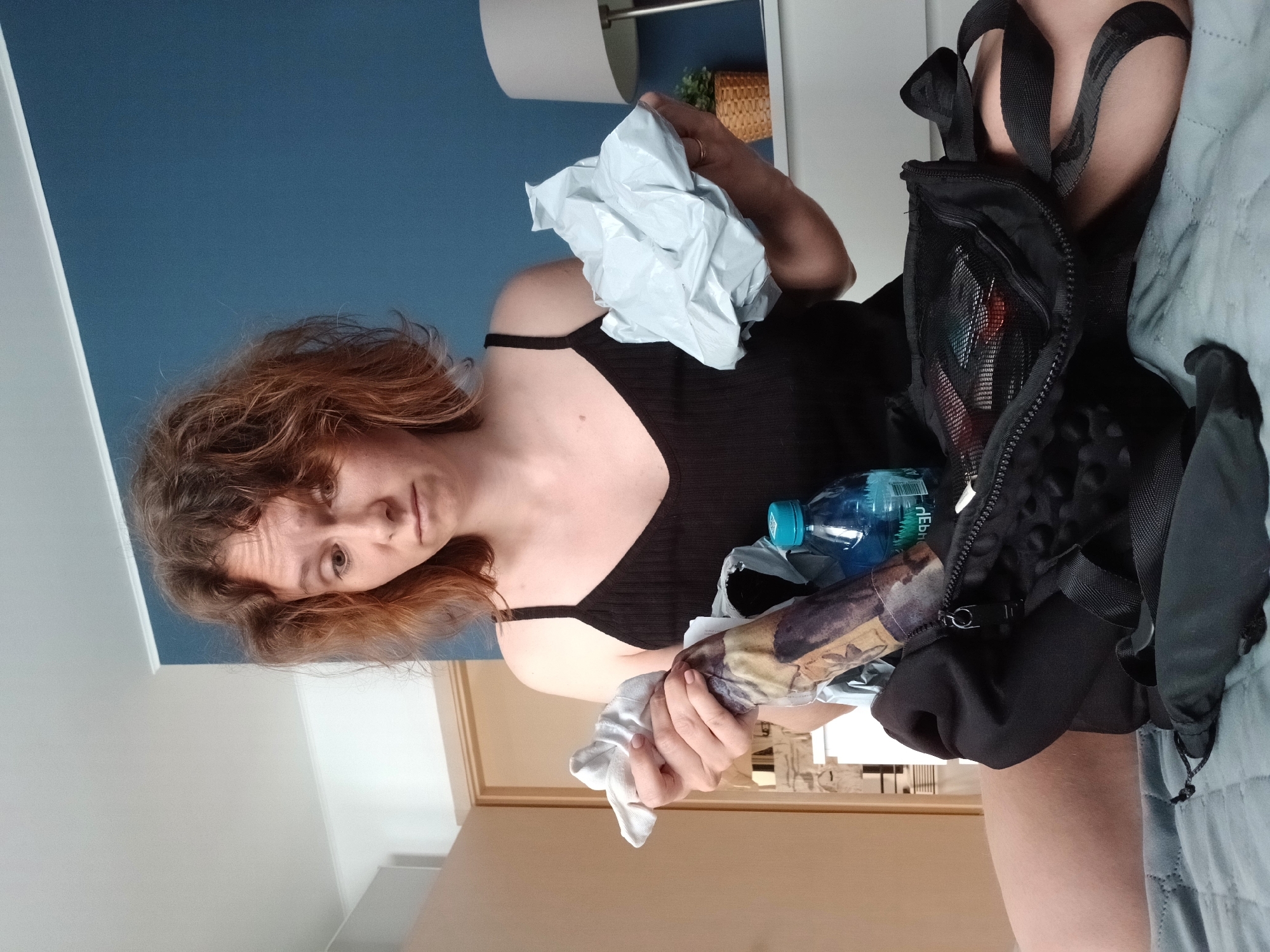 A young woman with medium-length, light brown, curly hair is sitting on a bed, looking down with a perplexed expression. She is wearing a black tank top. In her left hand, she holds a crumpled white piece of fabric. In her right hand, she is holding a light gray, crumpled object. In front of her, a black backpack is open, revealing various items inside, including a blue water bottle with a label that appears to have Cyrillic script, and other miscellaneous objects. The woman's legs are visible, and she is sitting on a gray quilted blanket. The background shows a blue wall, a doorway, and a white dresser with a lamp and a potted plant on it. The overall scene suggests a moment of packing or organizing, with the woman appearing uncertain about the items she holds. The location is not specified in the image.
