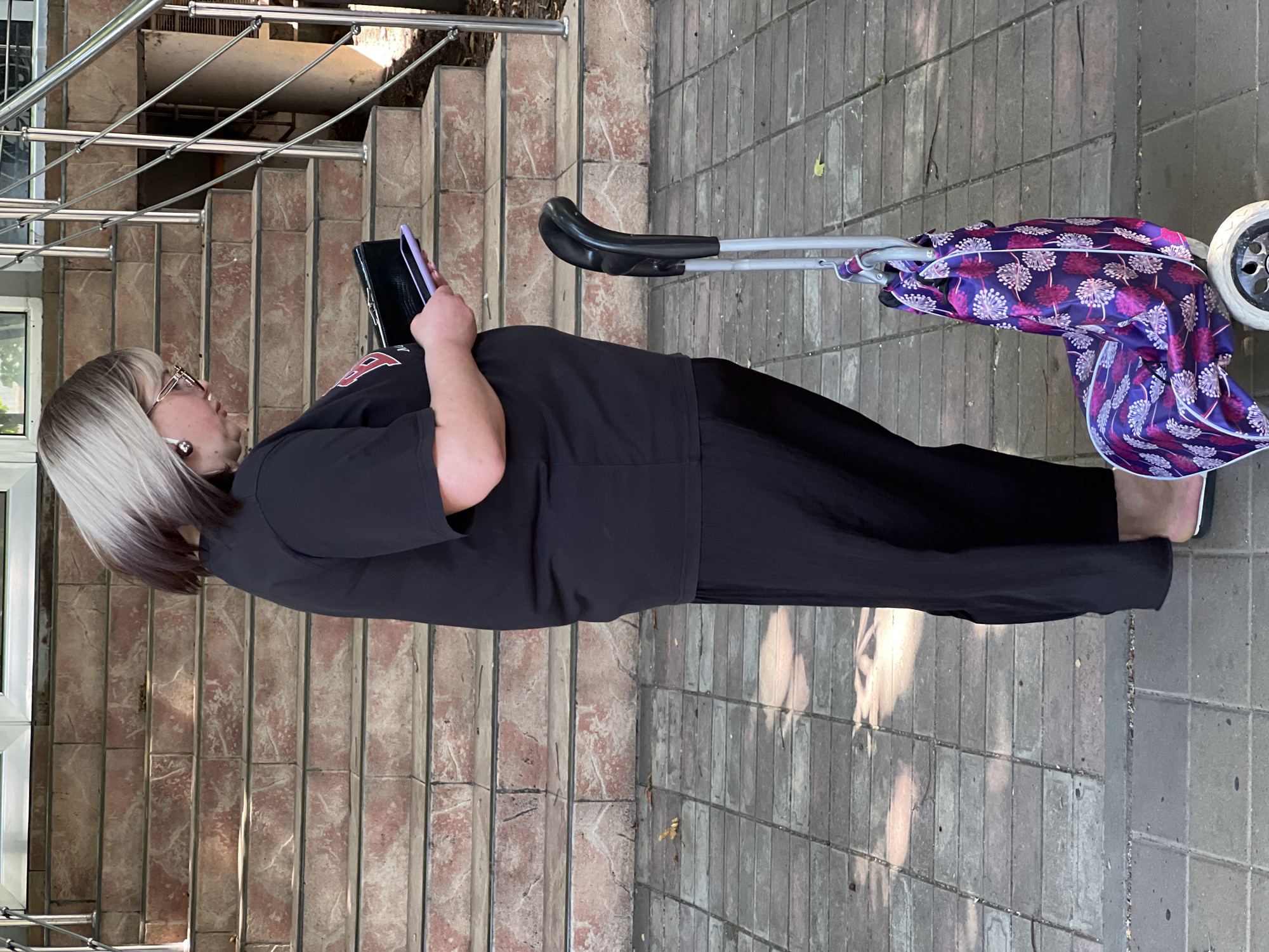 A person is standing on a paved walkway next to a set of concrete stairs with pinkish-red treads. The person is wearing a black t-shirt and loose black pants, and is holding a black wallet and a purple smartphone. A shopping cart with a large purple bag adorned with white and pink floral patterns is positioned next to the person's right leg. The background shows a building entrance with a metal railing along the stairs. The image appears to have been taken in Krasnodar, Russia, based on the surrounding architecture and the time of year indicated by the foliage.