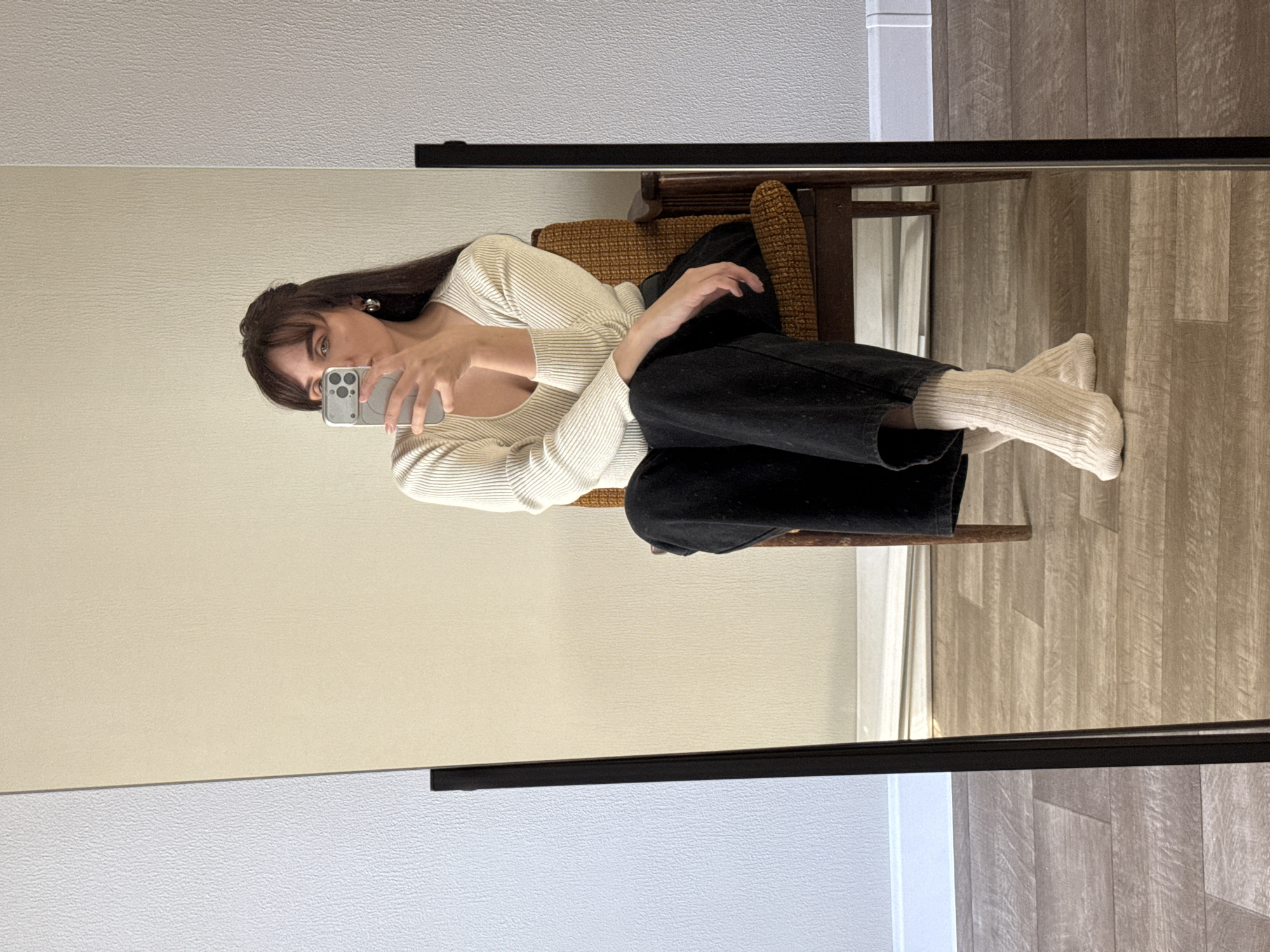 A young woman is sitting on a chair in front of a mirror, taking a selfie with her mobile phone. She is wearing a white, ribbed, long-sleeved top, black pants, and white socks. The chair is brown with a woven cushion. The floor is covered with light-colored wood-look planks. The background consists of a beige wall. The location is not discernable from the provided information. It can only be said that the photo was taken indoors.