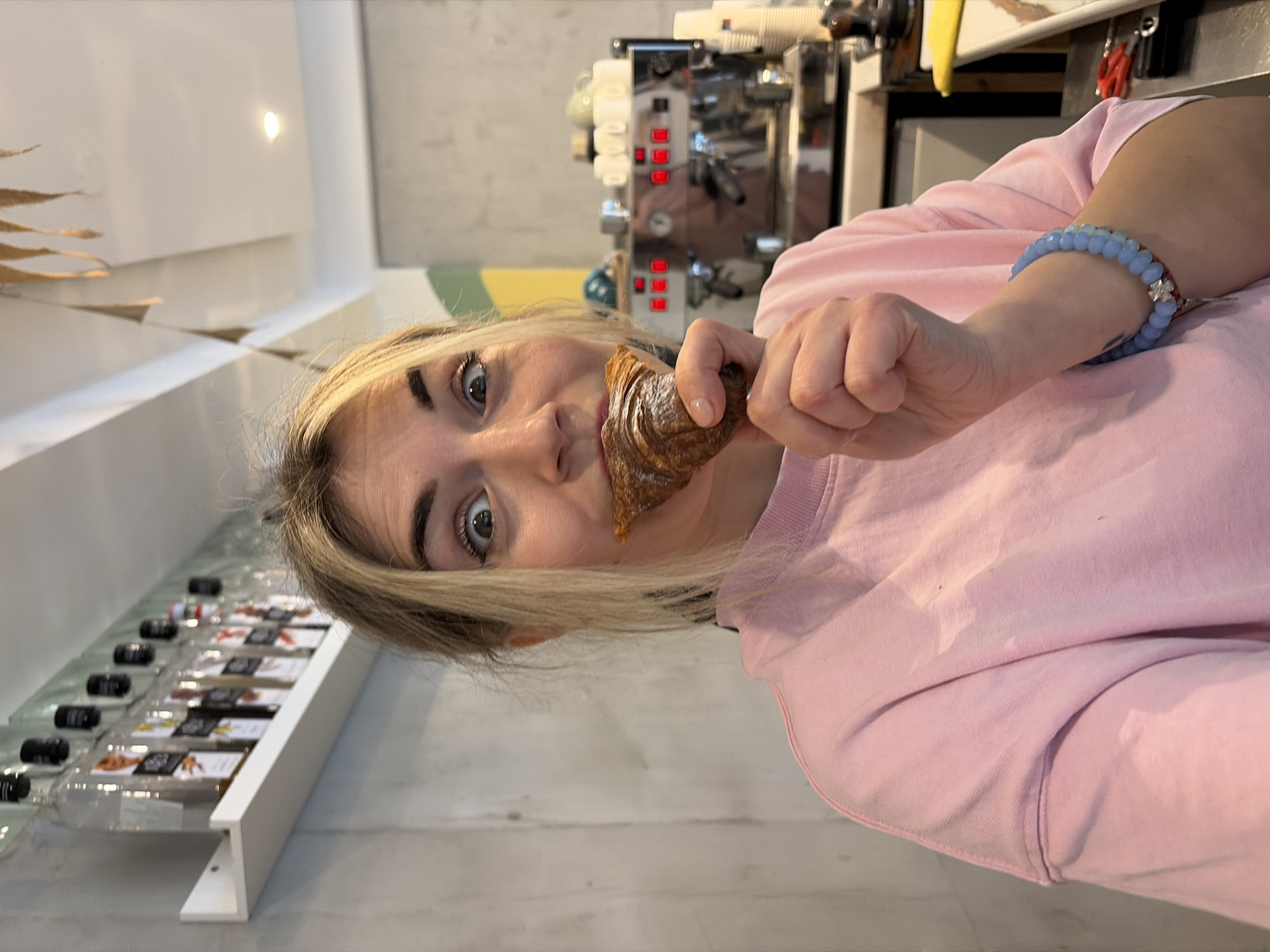 An indoor scene within a commercial establishment in Budva, Montenegro, likely a cafe, features a light-haired female subject from the chest up. She is looking directly at the camera with wide eyes and an open mouth, holding a partially consumed, golden-brown croissant in her right hand to her mouth. She wears a light pink long-sleeved sweatshirt and a light blue beaded bracelet with a silver-colored cross charm on her right wrist. The background includes a white shelf mounted on a concrete-textured wall to the upper left, displaying multiple clear glass bottles with black caps. To the right, a chrome commercial espresso machine with illuminated red indicator lights is visible, accompanied by stacks of white ceramic cups and disposable paper cups. The ceiling is white with a recessed light fixture and a decorative garland of brown, leaf-shaped elements. A section of the wall on the right displays a curved design painted in green and yellow/orange. The specific location within Budva cannot be determined from the visual evidence.
