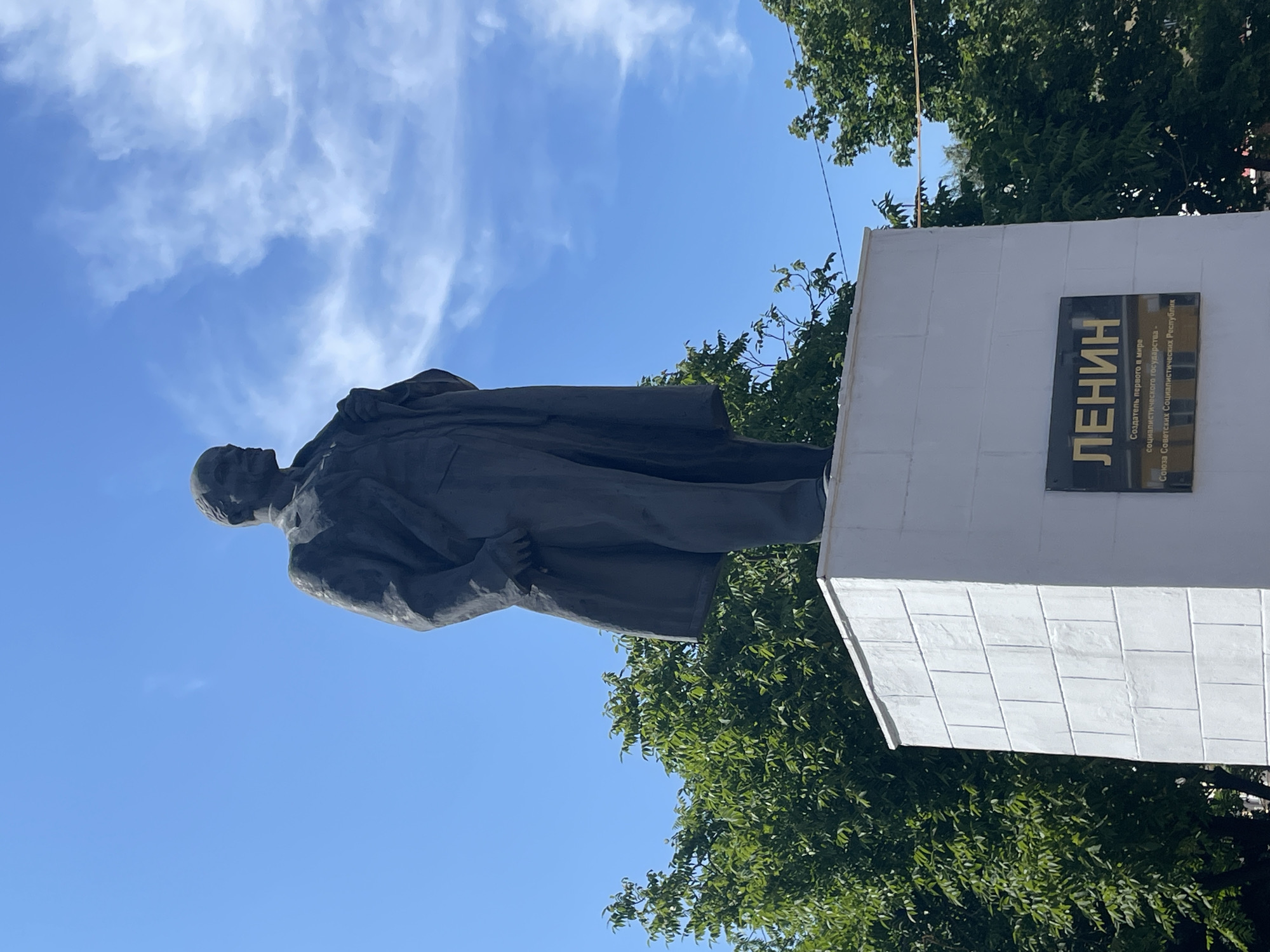 A full shot captures a dark, bronze statue of Vladimir Lenin standing on a tall, white, rectangular pedestal in Zavolzhye, Russia. The statue faces slightly to the left, with Lenin's right hand tucked into his coat pocket and his left hand holding the lapel of his jacket. He is dressed in a suit and overcoat. The pedestal has a plaque on its front with Cyrillic lettering. The background consists of a bright blue sky with wispy white clouds and lush green trees on either side of the statue. The lighting suggests it is daytime, likely a sunny day.