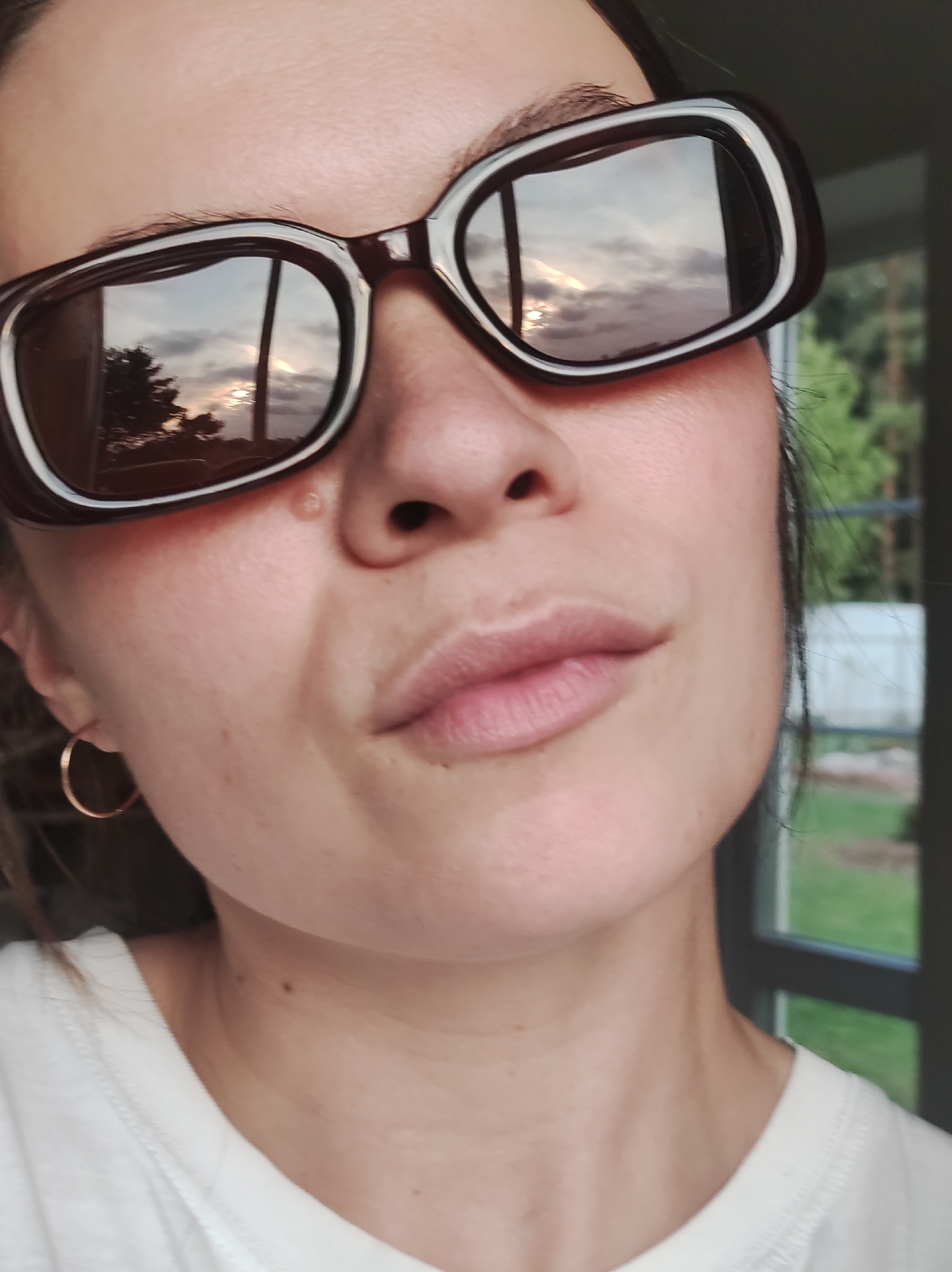 A close-up, slightly angled self-portrait shows a woman wearing sunglasses with dark frames. The reflection in the lenses reveals a sky with clouds and a setting sun, along with tree silhouettes. She wears a white t-shirt and small gold hoop earrings. There is a blemish near her nose. A window with a black frame is visible in the background, along with glimpses of green foliage. The photo's location is presumed to be in Фоки, Russia.