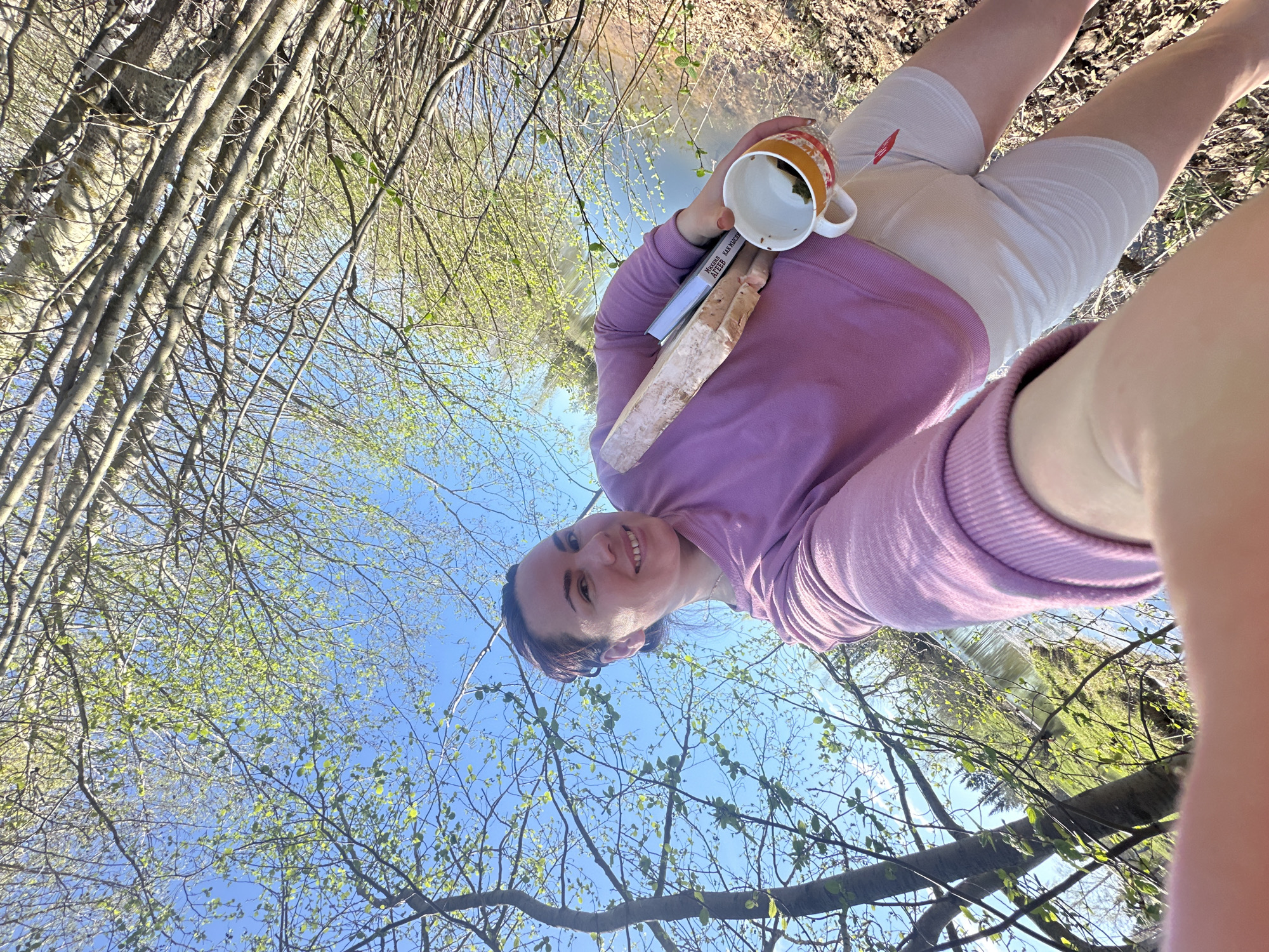 A young woman stands outdoors near a body of water, possibly a stream or river, surrounded by trees. The picture is taken from a low angle, looking up at the woman and the sky.
The woman is wearing a purple sweater and white shorts, with what appears to be a compression bandage on her thighs. She is holding a mug in one hand and a book in the other. She has a slight smile on her face.
The environment is a natural setting with trees and a water feature. The trees have new leaves budding, suggesting it is spring. The sky is blue and clear, with branches and leaves visible against it.
Based on the location provided, Черёмушки, Russia, this could be a park or wooded area within the city or on its outskirts.