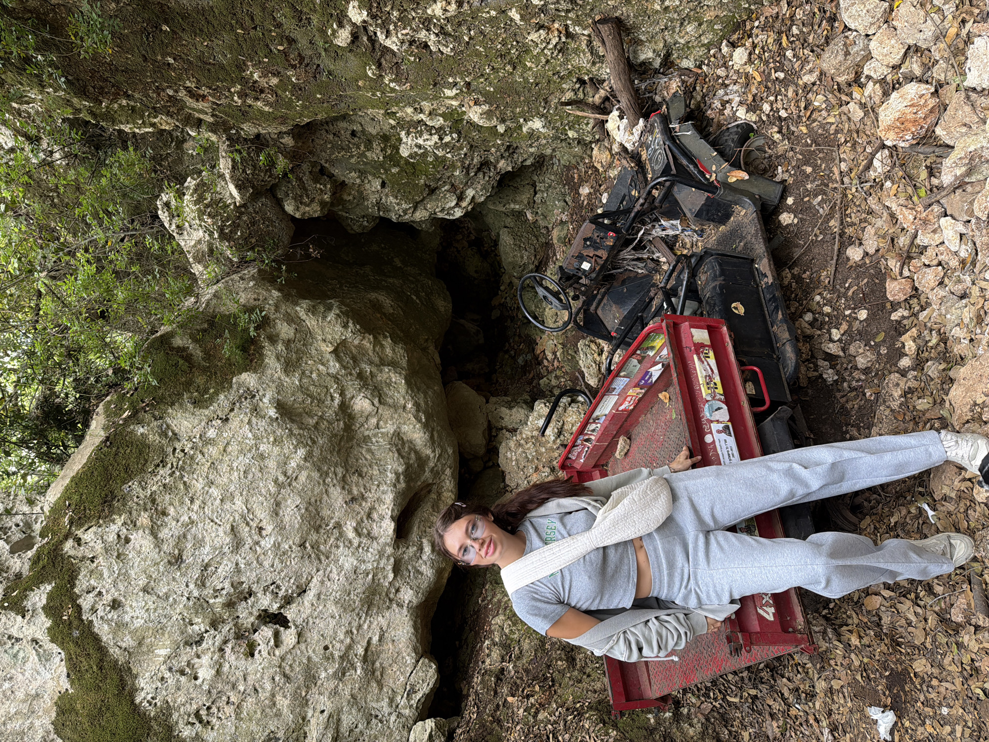 A young woman lies on her back on a red utility trailer. She is wearing a gray sweat suit, sneakers, and light pink sunglasses. A white purse rests on her hip. The utility trailer is next to a black utility vehicle. Behind the vehicle is a small cave. The ground is covered with rocks and leaves. The location is in a natural area, possibly a park, within the city of Tirat Karmel, Israel.
