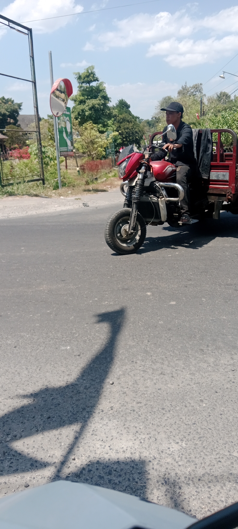A person wearing a black shirt and a black baseball cap is riding a red three-wheeled motorcycle. The motorcycle has a metal rack at the back, and a black garment is draped over the side of the rack. The motorcycle is traveling on an asphalt road. The scene appears to be in a semi-urban or suburban area, with trees and some buildings visible in the background. A convex mirror is mounted on a pole on the side of the road, and a billboard is partially visible behind it. The sky is blue with some white clouds. The time of day appears to be daytime, given the bright sunlight and the shadow cast by the motorcycle and rider. Based on the provided context, the location is Maguwoharjo, Indonesia.