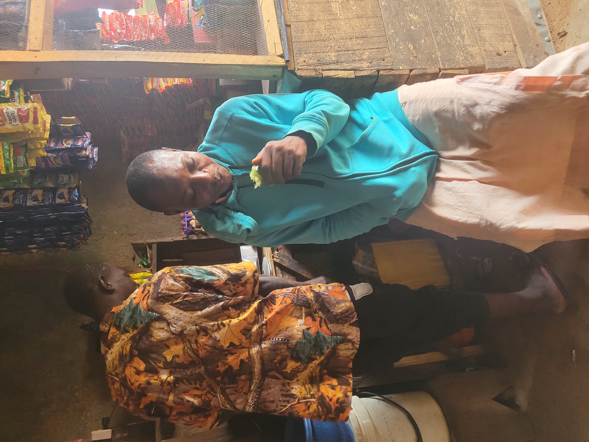 In Madalla, Nigeria, two individuals are present within a small, dimly lit shop. The person on the left, viewed from behind, wears a long-sleeved shirt with a camouflage pattern featuring autumn leaves in shades of brown, orange, and green. They are standing, facing away from the camera, with their back to the viewer. Their legs are clad in black pants.
To the right, a second person is seated, facing the viewer and slightly downward, holding a piece of green food in their right hand, which they appear to be eating or examining. This individual is wearing a bright teal hooded jacket with the zipper partially open and light pink fabric draped over their lower body, suggesting a skirt or loose trousers.
The shop's interior is rustic, with wooden shelves overhead displaying numerous packaged goods, including colorful bags of snacks and beverages. The walls appear to be rough plaster. To the right of the seated person, a section of the shop's counter or structure is visible, constructed from weathered wooden planks and covered with a wire mesh screen, behind which more merchandise is stacked. On the left side of the image, a large white bucket and a blue container are partially visible. The floor is dirt or concrete, showing signs of wear. The lighting suggests natural light entering from the front or side of the shop, casting shadows and highlighting dust particles in the air.