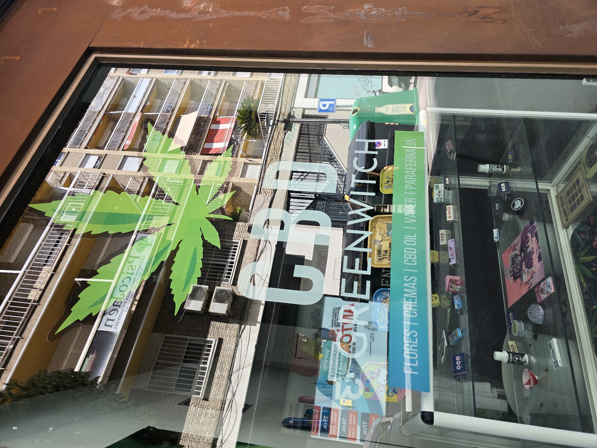 The image shows a shop window display for a CBD (cannabidiol) store named "GREENWITCH". The window features a large green cannabis leaf graphic and white lettering advertising "CBD GREENWITCH". Below this, a turquoise banner lists "FLORES | CREMAS | CBD OIL | VAPER | PARAPHERNALIA" in white text.
Inside the display cases, various products are visible, including small boxes, rolling papers, grinders, bottles, and packaging for CBD products. Some items are branded with "OCB".
The reflection on the window shows a building across the street with multiple balconies and awnings, some of which are red and white striped. There are also air conditioning units mounted on the building's facade. A blue parking sign is visible in the reflection.
The shop appears to be located in the city of Jaén, Spain, as indicated by a partially visible sign in the reflection with text that appears to be in Spanish. Specifically, the text on the building in the reflection includes "PSICOLOGÍA" and what looks like "CENTRO DE PSICOLOGÍA Y LOGOPEDIA", which translates to "Psychology Center and Speech Therapy Center". This type of establishment is often found in urban areas.