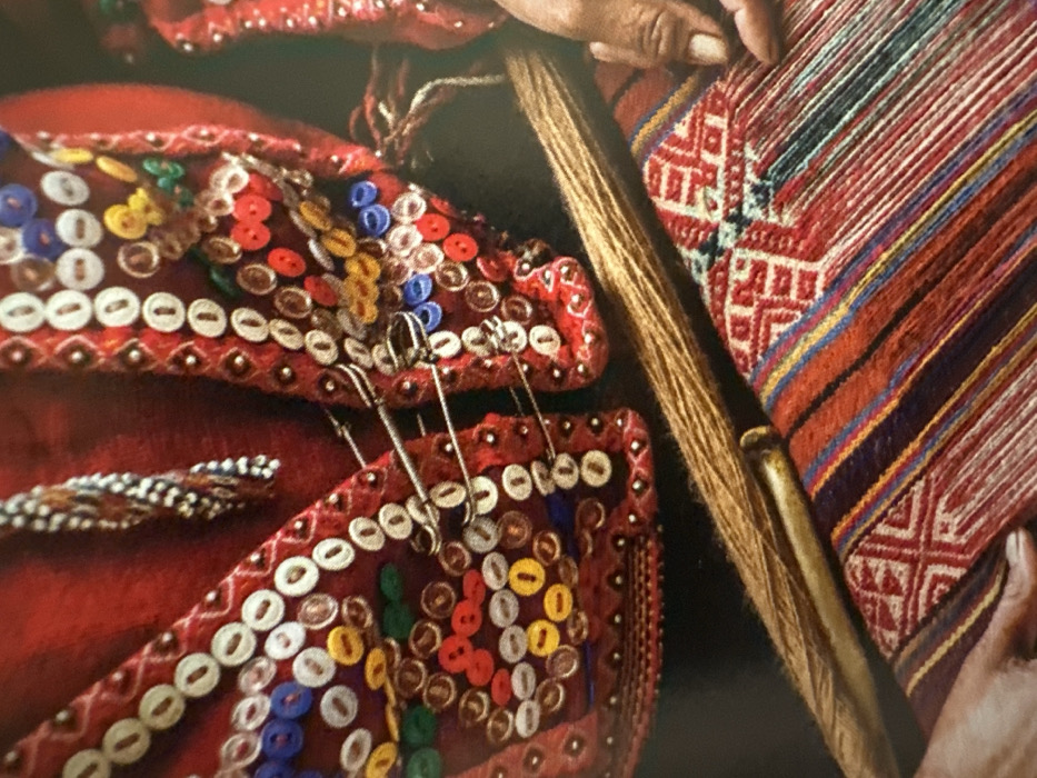 A close-up reveals handwoven textiles in vibrant hues of red, blue, and gold. The fabric features intricate geometric patterns. A red cloth is adorned with multicolored buttons and white shells, secured with safety pins. A spindle and hand are visible, suggesting the weaving process. The image highlights the artistry and detail in traditional textile crafts.