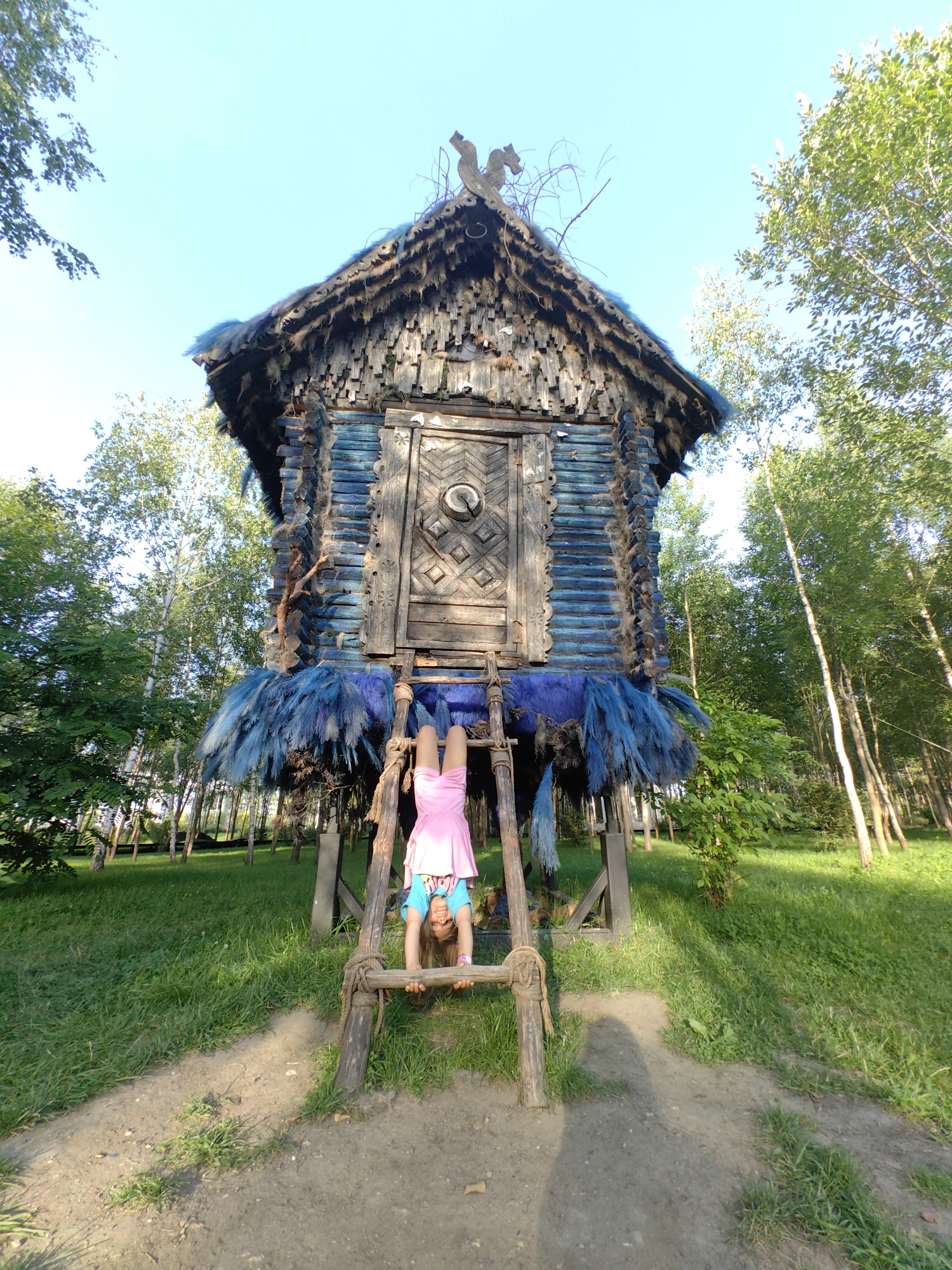 A young girl in a pink dress and blue shirt is doing a handstand on a wooden ladder in front of a rustic, wooden structure. The structure resembles a small, old-fashioned house or cabin, with a steeply pitched roof covered in what appears to be wooden shingles and adorned with fur or feathers. The walls of the structure are made of logs, some painted blue, and decorated with carved wooden elements and more blue fur or feathers. A wooden door with a circular decorative element is visible in the center of the structure. The ladder is made of rough-hewn wood and tied with rope.
The setting is outdoors, with lush green grass covering the ground and a dirt path leading towards the structure. Tall trees with green foliage surround the area, indicating a park or wooded environment. The sky is clear and blue, suggesting it is daytime with pleasant weather. The lighting indicates it is likely late afternoon, with the sun casting shadows on the ground. The girl's expression is not clearly visible due to the angle, but her pose suggests a playful or athletic activity. The overall atmosphere is whimsical and outdoorsy. The location is identified as Moscow, Russia. No visible text is present in the image.