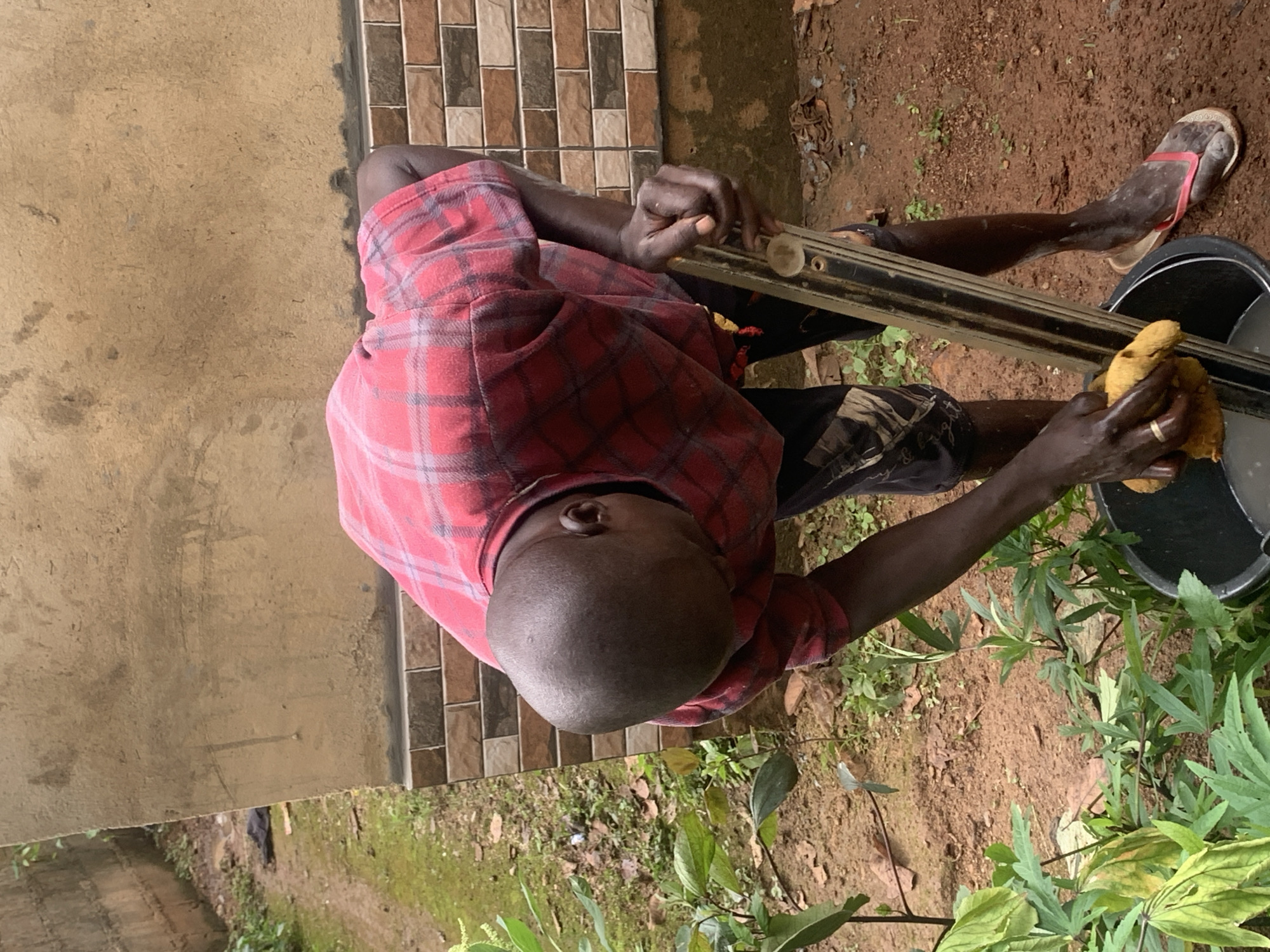 The image depicts a person wearing a red plaid shirt and dark shorts, stooped over a black bucket. The person is holding a yellow sponge in their right hand, which is submerged in the bucket's contents, likely water. In their left hand, the person is gripping a long, metallic tool with a clear level bubble. The bucket is positioned on a patch of dirt, with some green plants to the left. In the background, a textured wall, possibly made of concrete and adorned with a band of brick tiles, rises upwards. The ground is a mixture of red dirt and patches of green grass. The scene is located outdoors in a setting that appears to be a residential area in Agban, Nigeria.