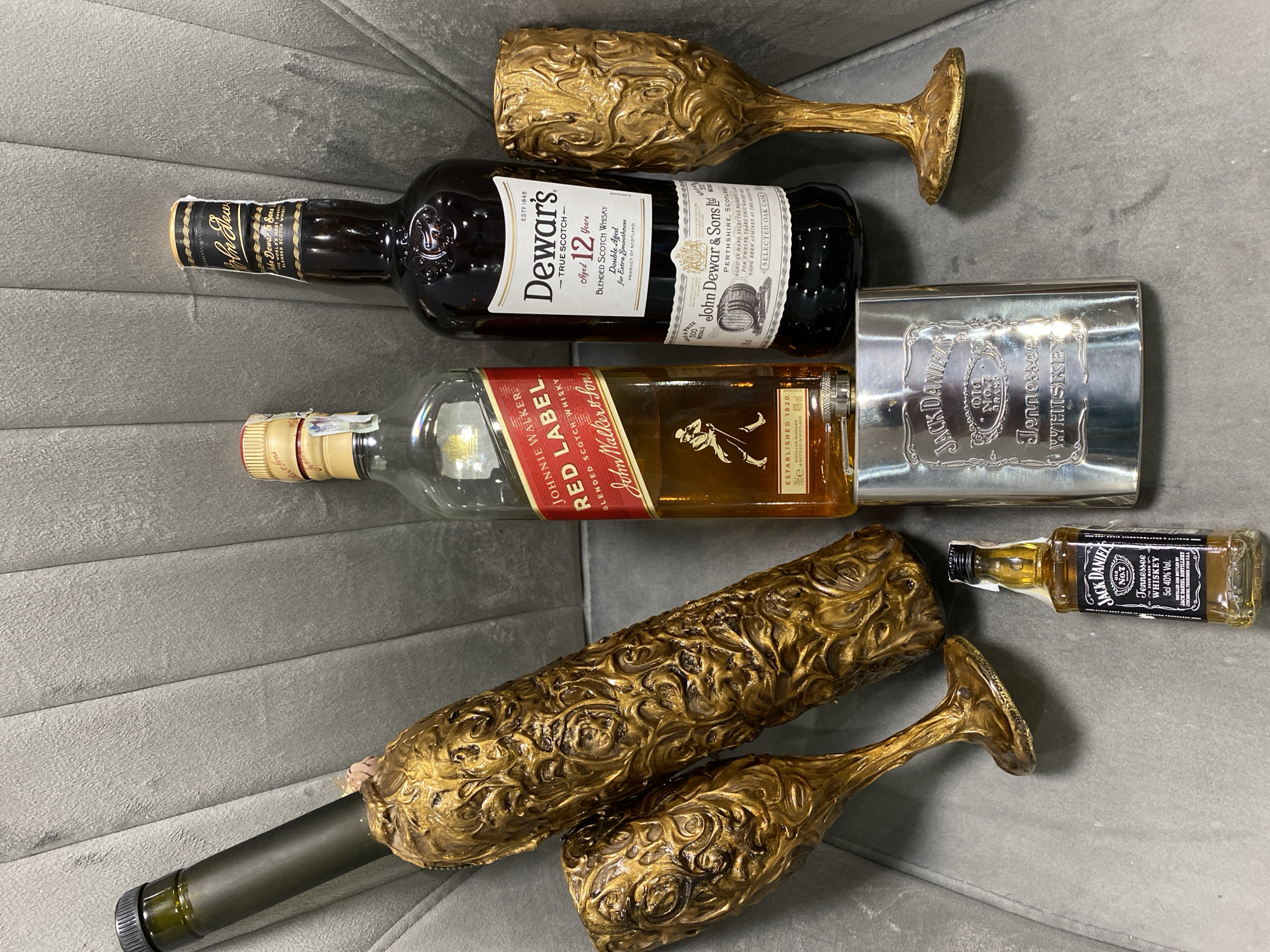 A flat lay arrangement of alcoholic beverages and glassware sits on a textured grey surface, possibly a sofa. The items include a tall, narrow bottle with a dark green neck and a decorative gold casing, two ornate gold goblets, a bottle of Johnnie Walker Red Label Scotch Whisky, a bottle of Dewar's 12 Year Old Blended Scotch Whisky, a small bottle of Jack Daniel's Tennessee Whiskey, and a silver hip flask embossed with the Jack Daniel's logo. The scene does not provide any information about its specific location within the city of Pleven, Bulgaria, or any details about people, actions, or interactions.