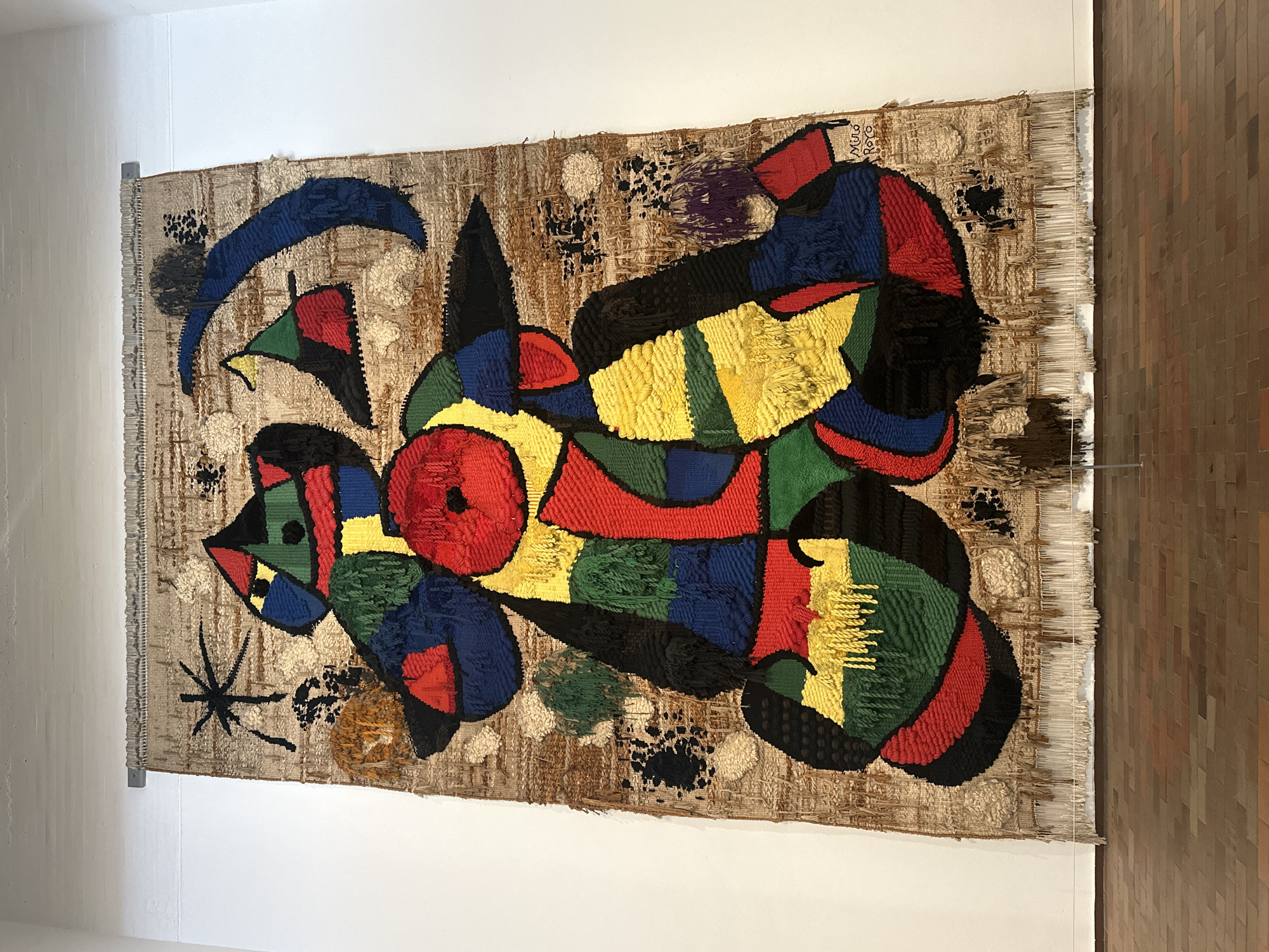 The image depicts a large, colorful tapestry hanging on a white wall. The tapestry features abstract shapes and figures in bold colors such as red, blue, yellow, green, and black, woven into a textured, neutral-toned background. The tapestry is hung using a metal rod and bracket system. The right side of the image shows a wall with a tiled surface of brown and orange tones. The scene appears to be indoors, likely in an art museum or gallery. Based on the style and subject matter, the tapestry is likely by Joan Miró, and the setting is potentially a museum in Barcelona, Spain dedicated to his work. The lack of people and action focuses the viewer’s attention on the tapestry as the main subject of the image.