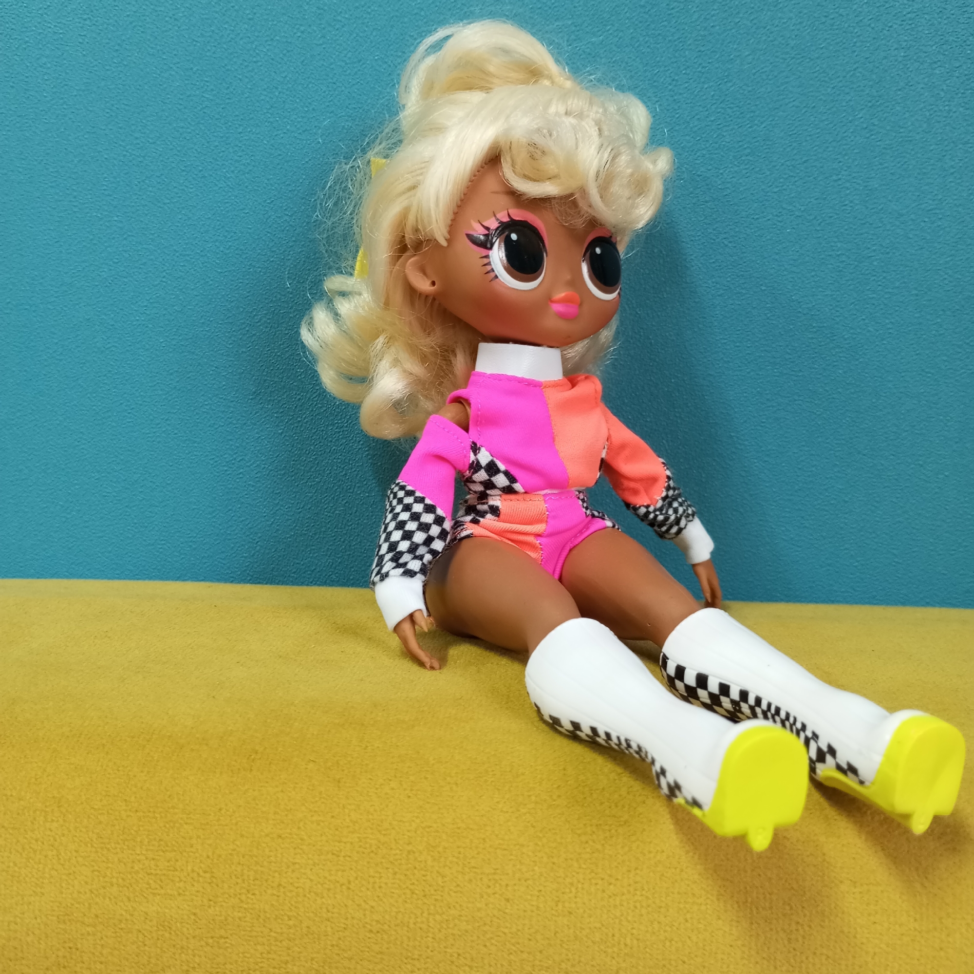 A doll with tan skin is sitting on a yellow surface against a turquoise wall. The doll has blonde, curly hair pulled up in a high ponytail with a yellow ribbon, and large, brown eyes with pink eyeshadow. She is wearing a pink and orange tracksuit with checkerboard sleeves and boots with yellow soles. The doll is sitting with her legs extended.
The location in the city of Pervomaysky, Russia is not discernible from the image.
