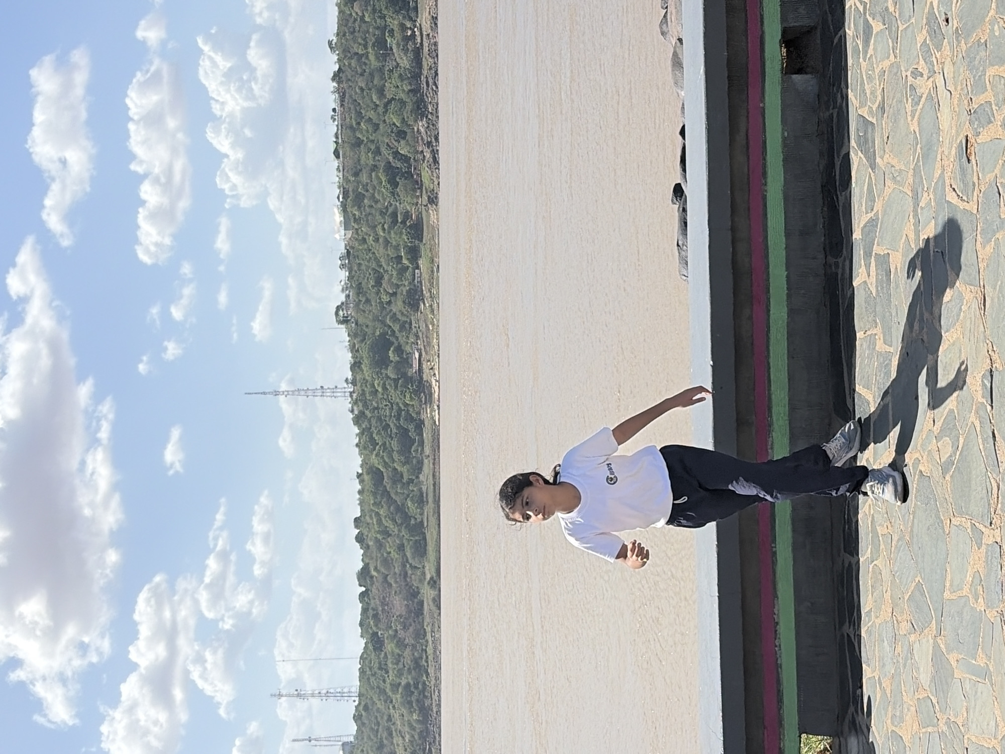 A full shot depicts a woman in a white t-shirt and black sweatpants standing on a tiled surface in Ciudad Bolivar, Venezuela. Behind her is a beige-colored river or expanse of water bordered by trees and a distant city skyline. The sky is blue with puffy white clouds. The woman is casting a shadow on the tiled surface. Behind her is a wall or ledge with painted vertical stripes of gray, dark red, and green. The image is rotated counter-clockwise by approximately 90 degrees.