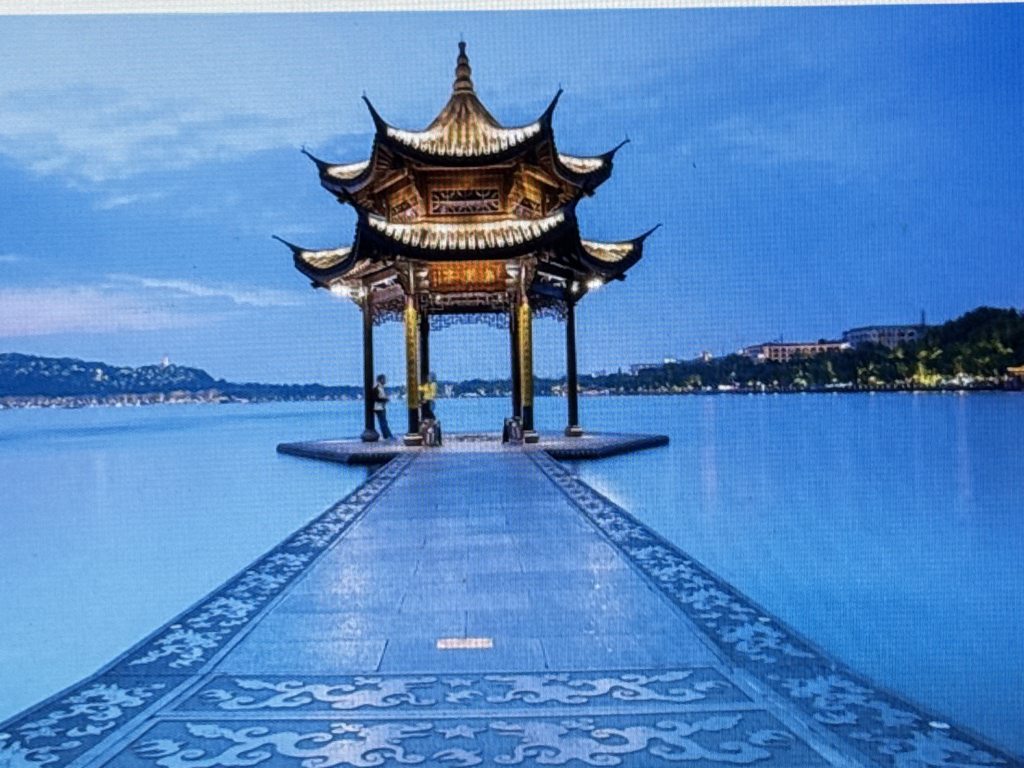 A wide-angle shot captures a traditional Chinese pavilion situated in the middle of a tranquil body of water. The pavilion, with its multi-tiered, ornate roof and golden accents, stands on a platform accessible by a long, stone walkway adorned with intricate cloud-like carvings. The walkway stretches towards the viewer, creating a strong sense of perspective. Two figures are visible on the platform, one standing near the pavilion's entrance. The water reflects the twilight sky, which transitions from a deep blue to a lighter hue at the horizon, where a distant cityscape with buildings and trees is faintly visible. The overall atmosphere is serene and majestic.