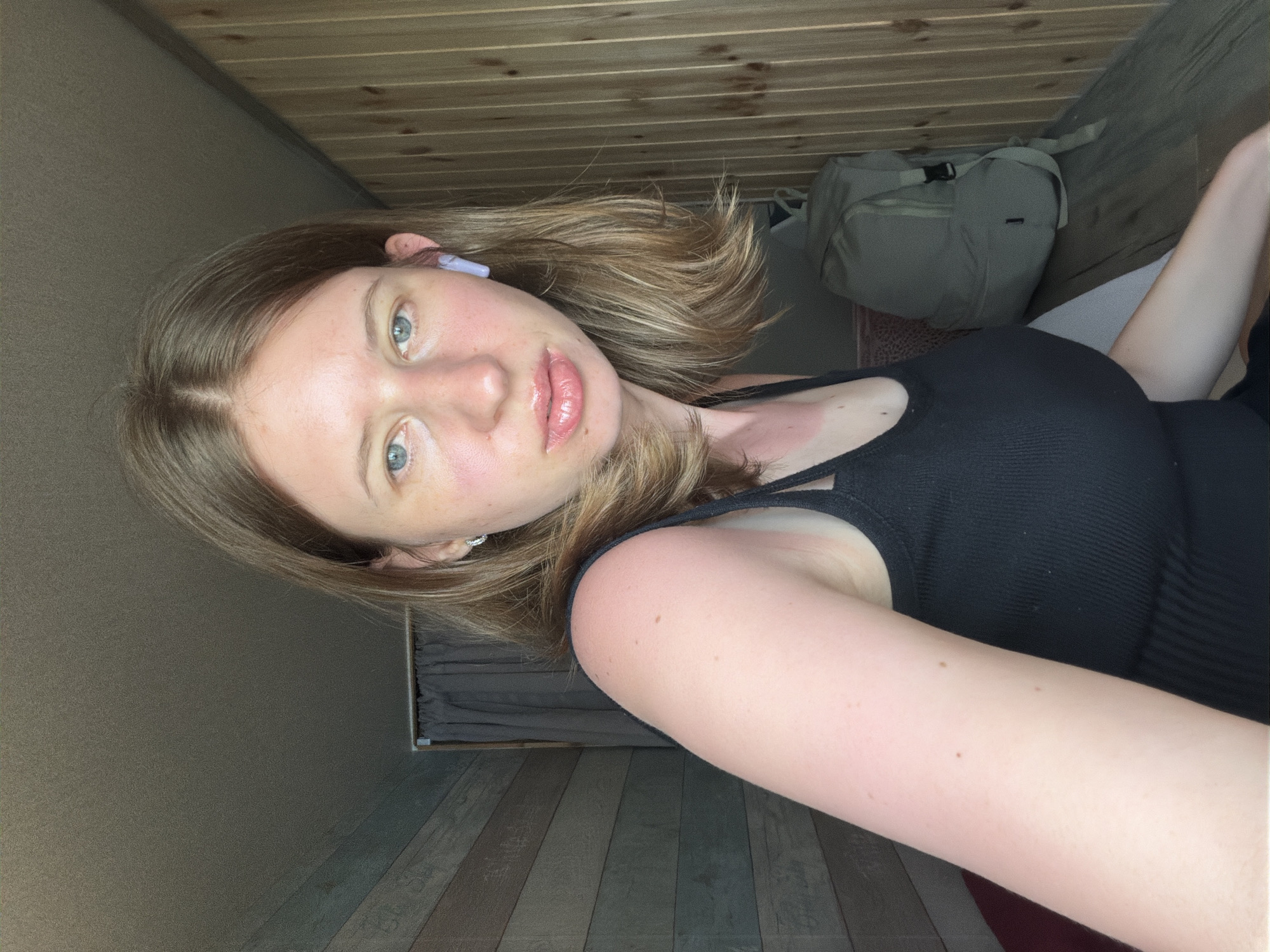 A young Caucasian woman with shoulder-length blonde hair and blue eyes is taking a selfie in what appears to be a room with wooden walls and a ceiling. She is wearing a black tank top and a light purple wireless earbud in her right ear. There is a pale green backpack behind her, leaning against the wall. Her skin appears lightly tanned and sunburnt on her right arm and shoulder. The room is dimly lit, with natural light coming from an unseen source. The image captures her from the chest up, with her head turned slightly to her left. The background consists of wooden paneling on the right side and a darker, textured wall on the left, with a grey curtain visible in the upper left corner. There is no indication of the city of Gangneung-si, South Korea, in the image.