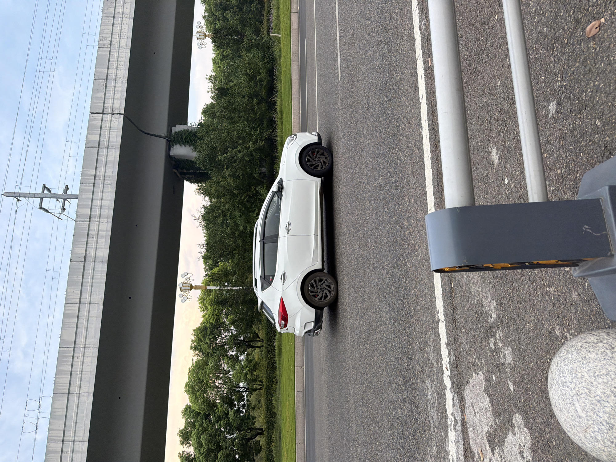 A white crossover vehicle drives on an asphalt road under an elevated structure. The car is white with black rims and is positioned in the middle of the frame, facing left. The elevated structure appears to be a highway overpass or bridge made of concrete, with visible support columns and overhead wires. Behind the road, there is a line of green trees and vegetation. The sky is overcast with some light visible. In the foreground, part of a grey barrier and a spherical object are visible. The scene suggests an urban or suburban setting, likely in Jiangjin, China, given the context provided.