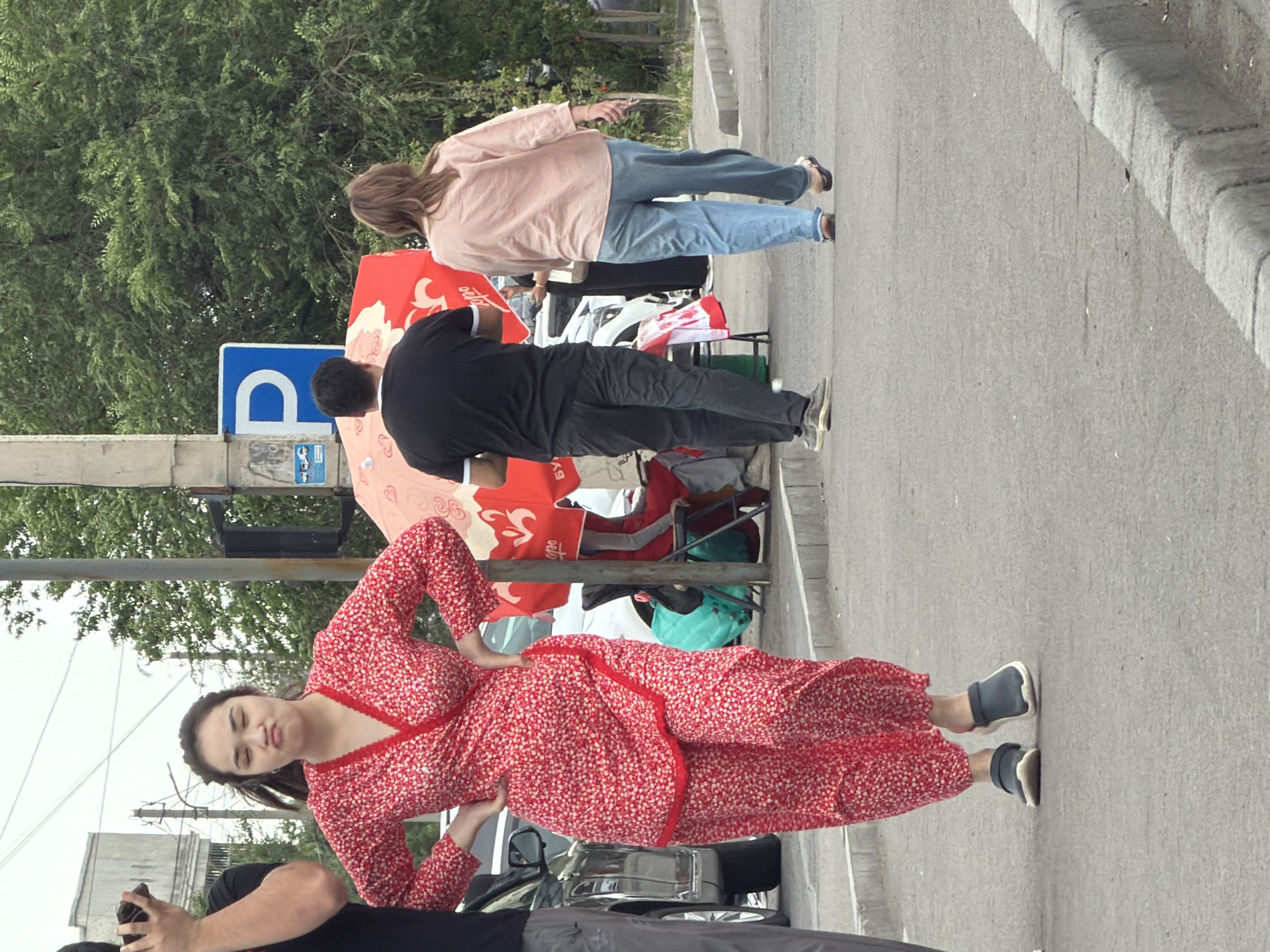 A young woman in a red floral print outfit with a v-neck top and wide-leg pants is standing with her hands on her hips, making a pouting face. She is on a paved street with a curb and sidewalk. Behind her, a man in a black t-shirt and grey pants is walking away from the camera. To his right, a woman in a light pink long-sleeved shirt and light blue jeans is walking away. In the background, there is a red umbrella, trees, a utility pole with a blue parking sign, and parked cars.