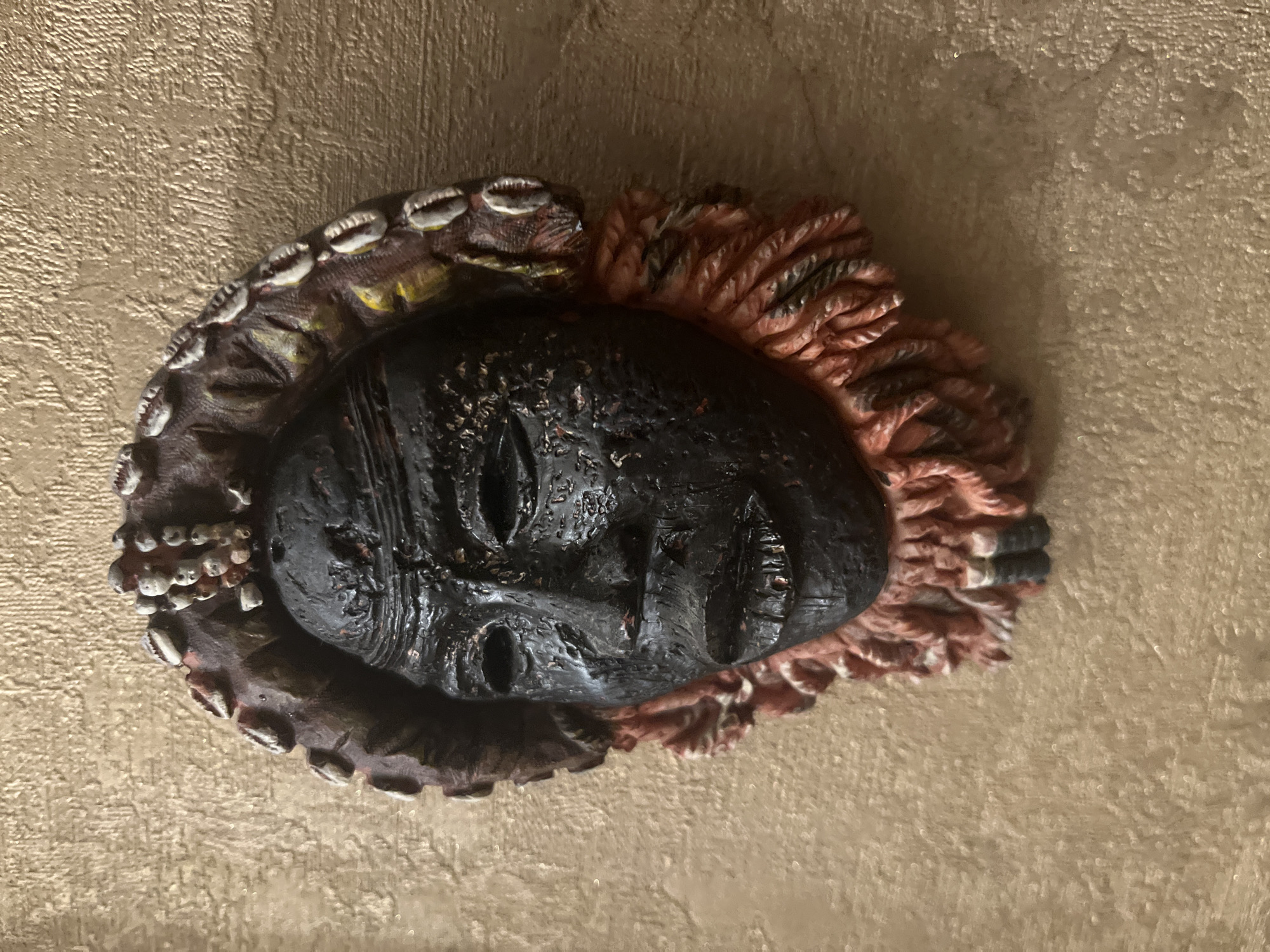 A close-up, eye-level shot shows a dark, intricately carved African mask mounted on a textured, light brown wall. The mask has a somber expression with deep-set eyes and full lips. It is adorned with a headdress that appears to be made of shells and beads, and a fringe of reddish-orange and black threads encircles the bottom of the mask. The wall has a rough, stucco-like texture and is illuminated from the side, casting a subtle shadow to the right of the mask. The image is focused on the mask, with the background wall slightly blurred. The specific location in Vladimirovka, Russia, is not discernible from the visual information provided.