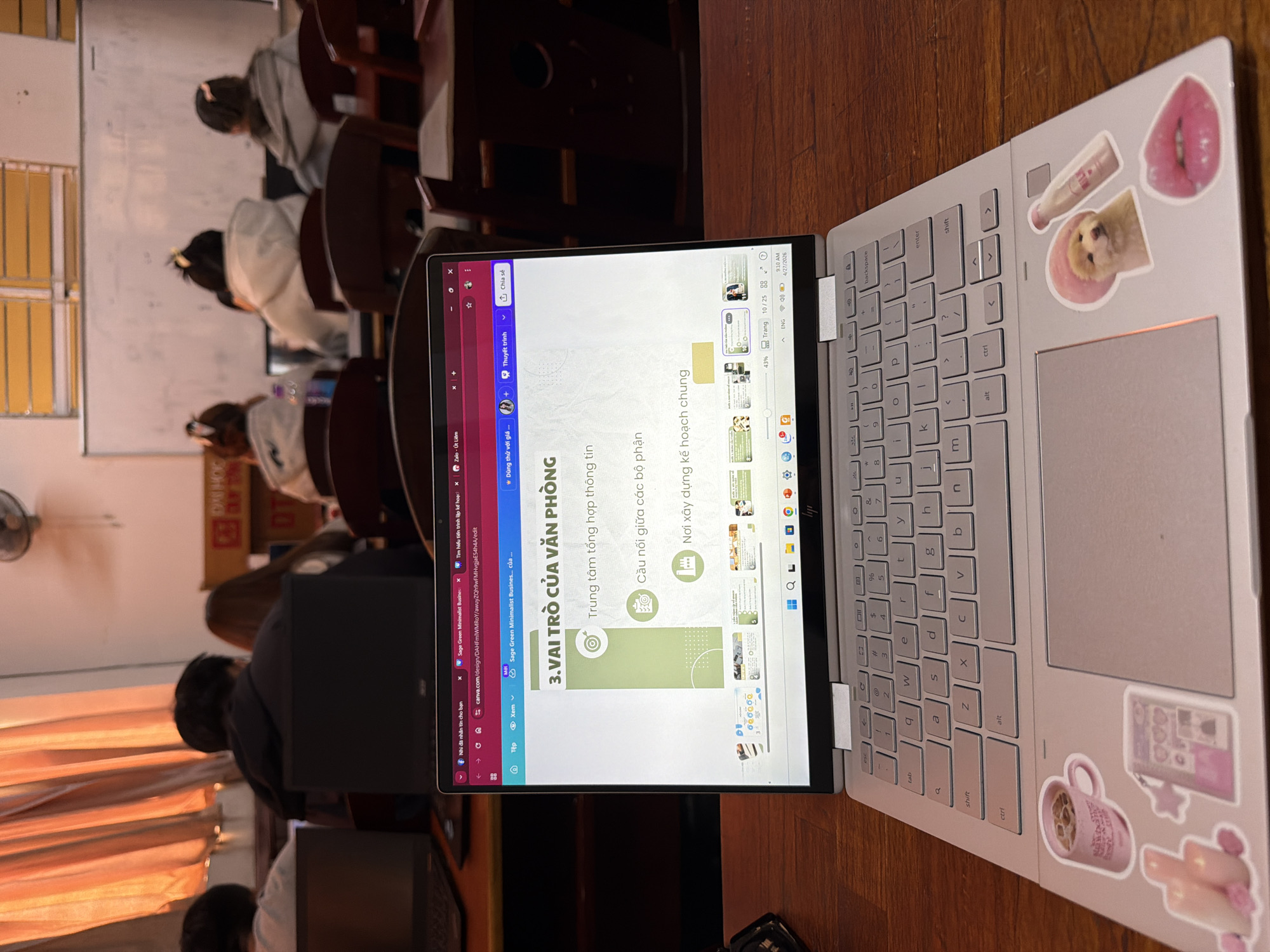 The image shows a laptop with an open screen displaying a presentation in Vietnamese. The title on the screen reads "3. VAI TRÒ CỦA VĂN PHÒNG" ("3. THE ROLE OF THE OFFICE"). The laptop has stickers on its outer surface, including images of lips, a dog, and a coffee mug. The laptop rests on a wooden surface. In the background, there are students seated in chairs, facing away from the camera, in what appears to be a classroom or lecture hall. The location appears to be in a city in Vietnam, given the Vietnamese text and possibly Đà Nẵng, Vietnam, but the image provides no information on what city the picture was taken in.