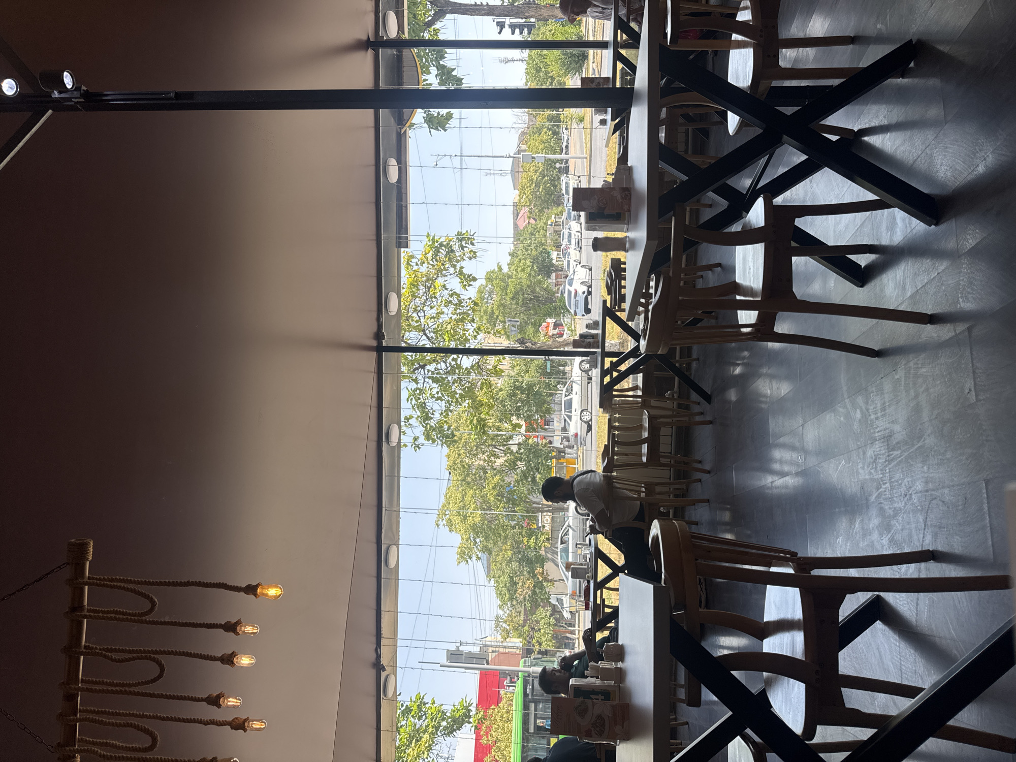 The photograph captures an interior view of a cafe or restaurant located in Shymkent, Kazakhstan, looking out through a large window. The perspective is from within the establishment, with tables and chairs visible in the foreground. The tables are modern, with dark, crisscrossed metal bases and light-colored wooden tops. Several wooden chairs with curved backs are arranged around the tables.
Through the expansive window, a street scene unfolds. Trees line the sidewalk, their green foliage vibrant against a bright, clear sky. The street is moderately busy with traffic, including cars and what appears to be a green public bus. Buildings and utility poles are visible in the background, indicating an urban setting. The sunlight streams in, casting distinct shadows on the polished floor inside the establishment.
In the middle ground, within the cafe and visible through the window, several patrons are seated at tables. One man in a green shirt is seated at a table, appearing to drink from a glass. Another man, wearing a white t-shirt, is looking at his phone while seated. There are other individuals and groups of people blurred or partially visible, suggesting a typical daytime scene in a public eatery. A decorative rope-and-light fixture hangs from the ceiling, adding to the cafe's ambiance.