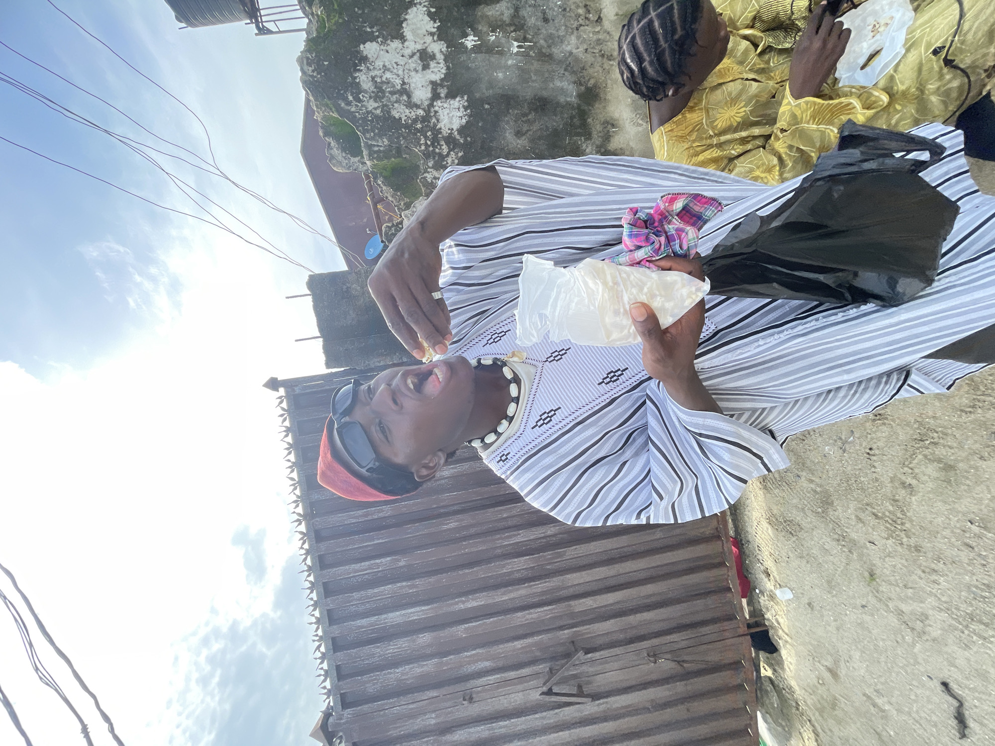 In Ado, Nigeria, a man in a striped robe, red cap, and sunglasses holds a plastic bag of food and eats from it while standing near a corrugated metal structure. Another person sits nearby wearing a gold-colored garment. Power lines crisscross the sky above.
