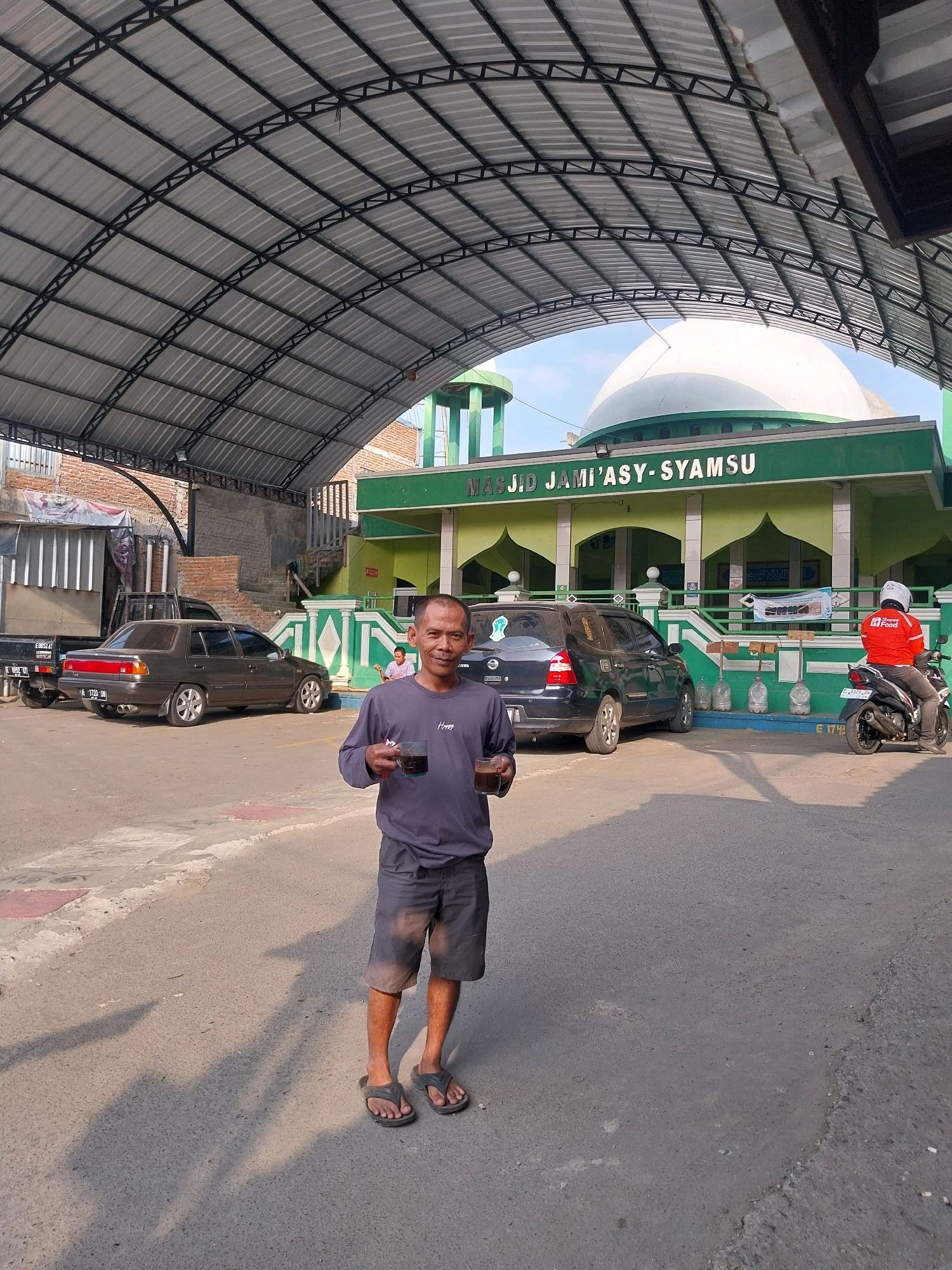 A man stands in front of a mosque in Cirebon, Indonesia. He is holding two clear plastic cups filled with a dark liquid. He is wearing a dark blue long-sleeved shirt and grey shorts, and appears to be smiling. Behind him, a mosque with a white dome and green accents is visible, with the words "MASJID JAMI'ASY-SYAMSU" written in white lettering above the entrance. Several cars are parked in the foreground, including a dark sedan and a black SUV. A motorcycle with a rider wearing a red jacket is parked to the right of the mosque. The scene is outdoors, under a metal canopy structure that covers a portion of the area. The ground is paved with asphalt.