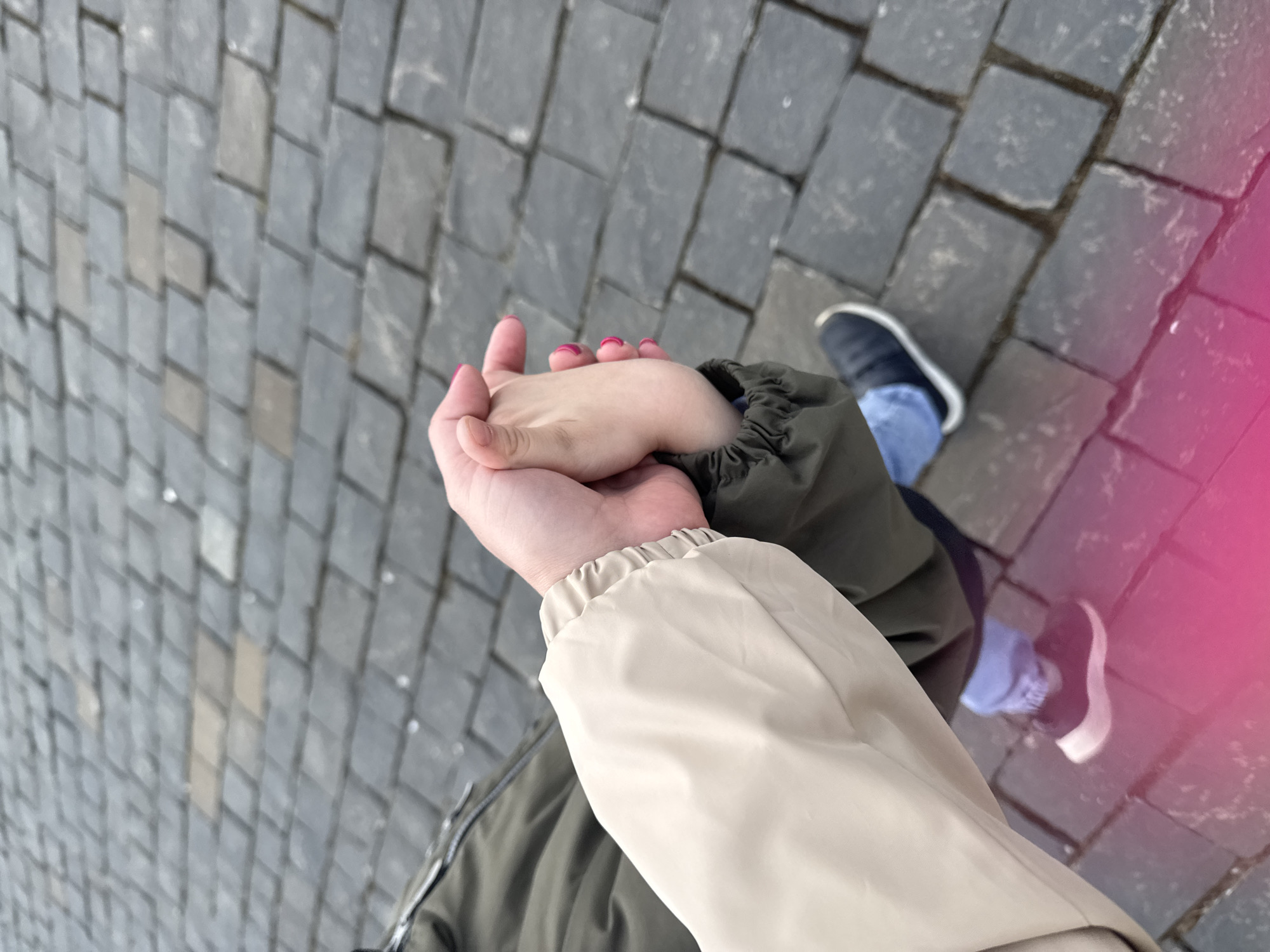 A high-angle shot shows two people holding hands while standing on a cobblestone street. One person wears a tan and green jacket and the other wears an olive-green jacket with blue jeans and black shoes. The cobblestones are gray and form a repeating pattern. A pink haze appears in the upper-right corner of the frame. The location is possibly Moscow, Russia.