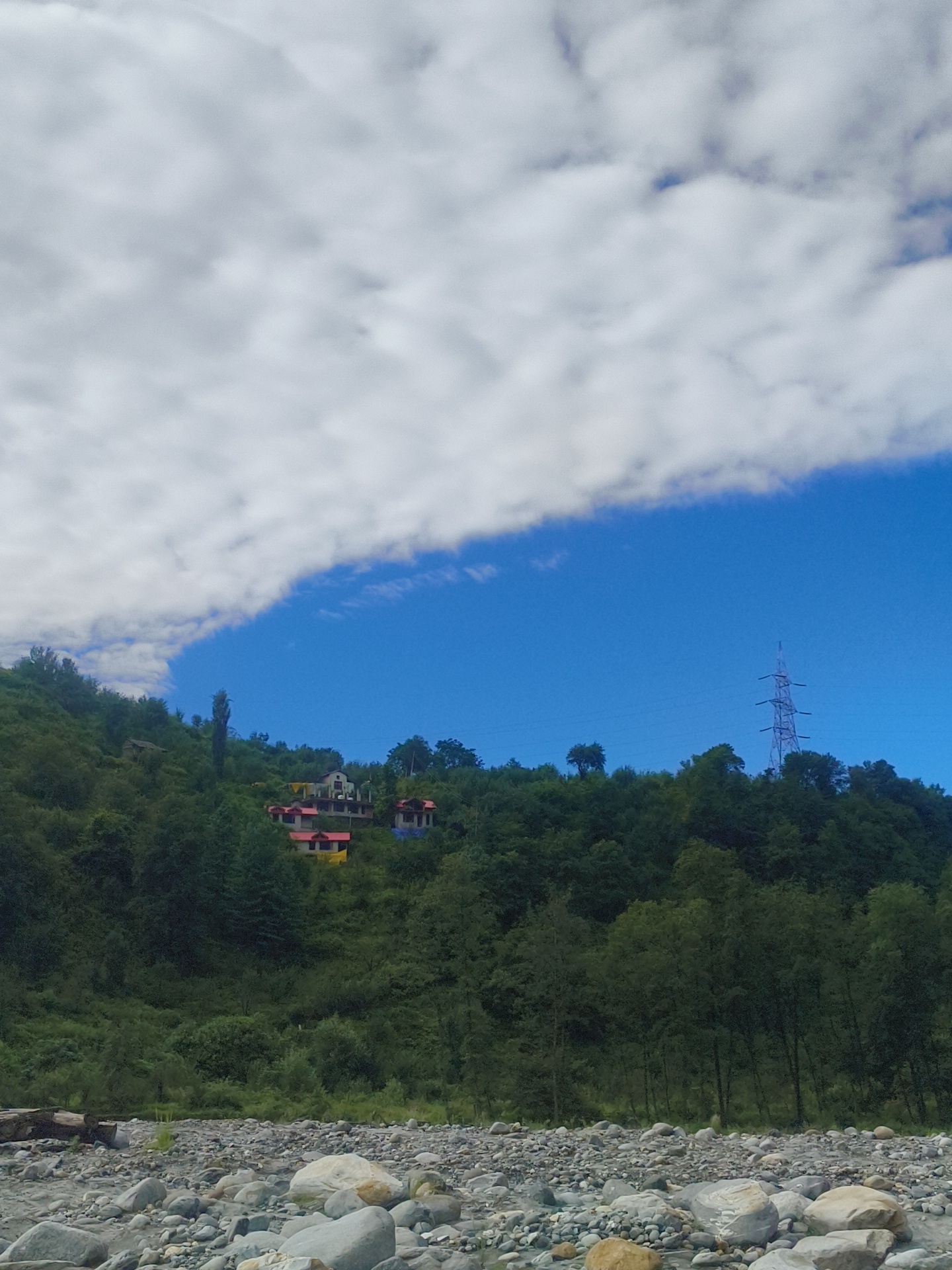 The image captures a serene landscape in Kalath, India. The foreground features a rocky riverbed with scattered stones of various sizes. Beyond the riverbed, a lush green hillside rises, densely covered with trees. Several houses with red roofs and yellow accents are visible nestled on the slope of the hill. In the background, a tall electricity pylon stands against a bright blue sky, partially obscured by large, white clouds. The overall scene depicts a tranquil natural setting with man-made structures integrated into the environment.