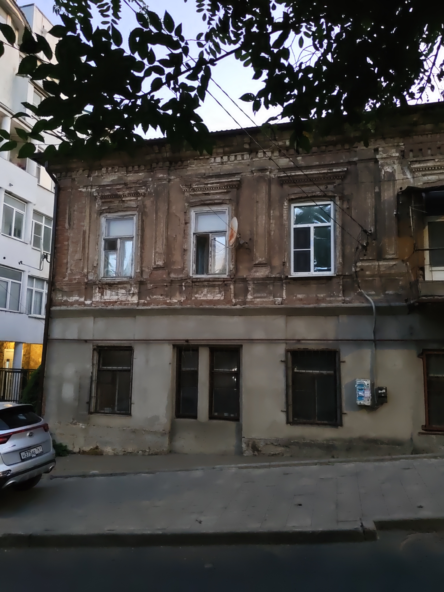 A silver Kia Sportage is parked on the street in front of an old, multi-story brick building with peeling paint. A modern, white building stands to the left of the older structure. The sky above is visible through the dark silhouettes of tree branches. The scene is located in Rostov-on-Don, Russia.