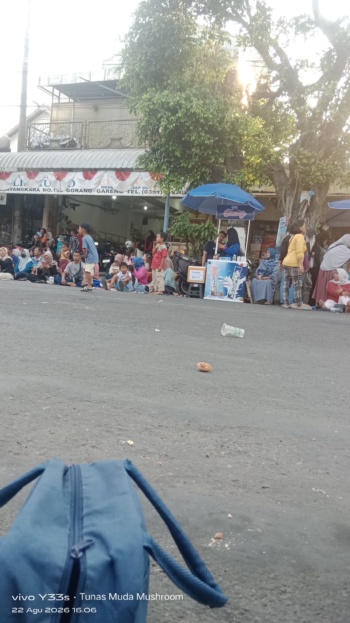 In Genengan, Indonesia, on a street lined with businesses, a diverse crowd of people, including men, women, and children, are gathered on the asphalt. Many are seated on the ground, facing a collection of stalls. One stall displays various drinks, some in bottles and others in cartons, beneath a blue umbrella. The atmosphere suggests a public event or gathering. A blue backpack lies in the foreground, its zipper partially open. The background reveals buildings, parked motorcycles, and lush green trees, with sunlight filtering through the foliage. The timestamp "22 Agu 2026 16.06" is visible at the bottom of the frame.