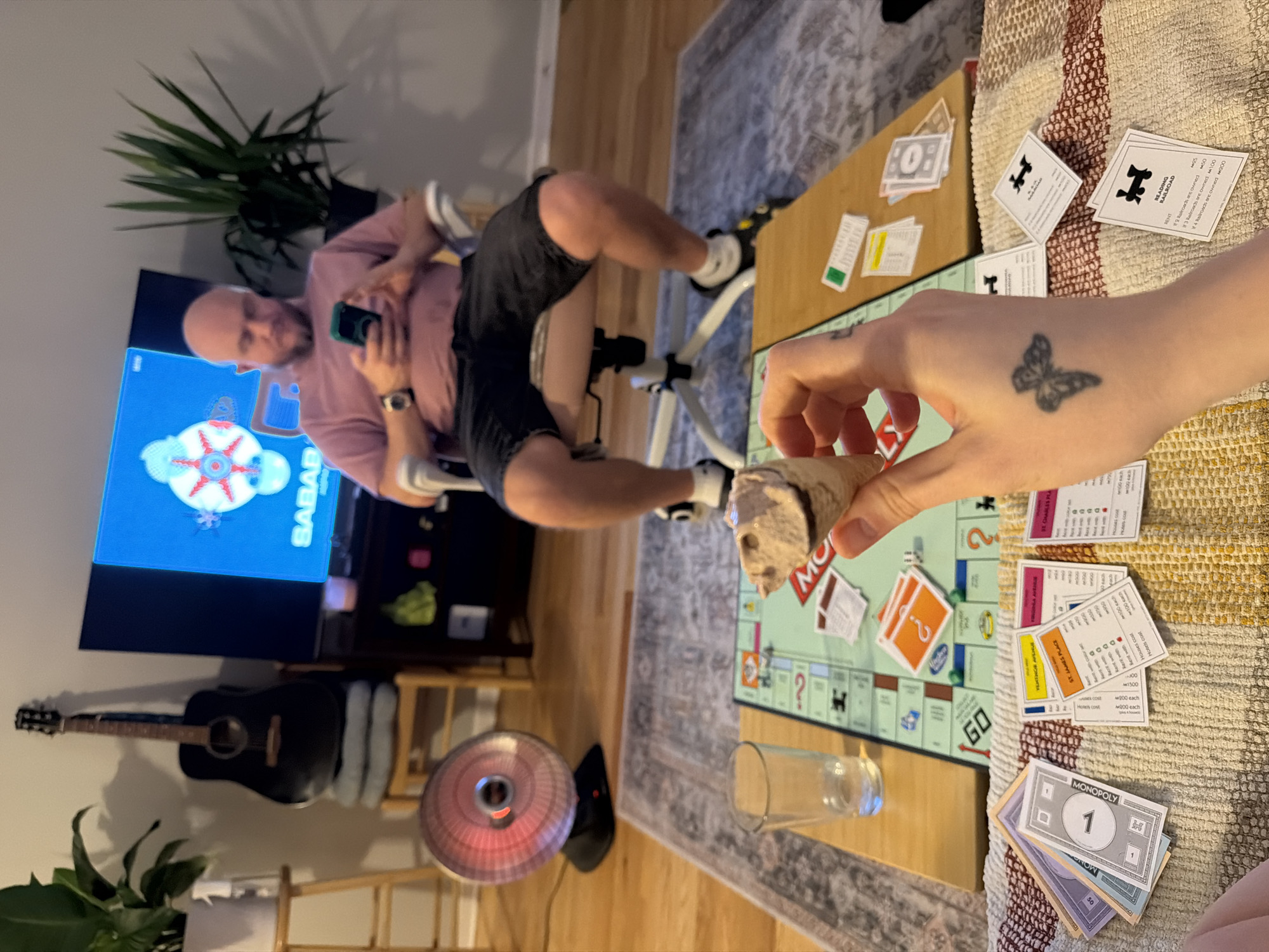 The image captures a casual indoor scene. A person sits in an exercise chair in front of a television displaying the word "SABAB" along with an abstract visual, and has a cell phone in their hands. A guitar leans against the wall next to the television, and there's a small space heater on the floor. In the foreground, another person is seated at a Monopoly board, with property cards and money visible. A stone sculpture of a face is being held over the board, seemingly acting as a playing piece. The scene is lit naturally, and a houseplant is situated in the background, suggesting a home environment. The image does not provide enough information to place the image within the specific city of Morton Grove, United States of America.
