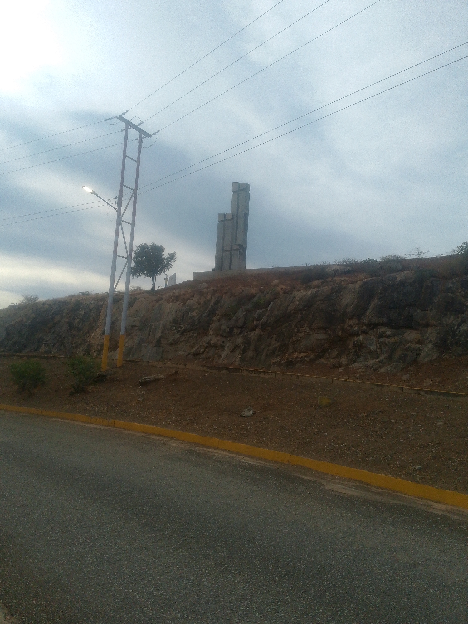 The image shows the monument to the heroes of independence, in Juan Griego, Venezuela. The monument consists of three tall concrete towers, on a hilltop. At the base of the hill is a road with a yellow curb. There's a utility pole with a streetlight near the road. Overhead, power lines stretch across the sky. The sky is overcast.