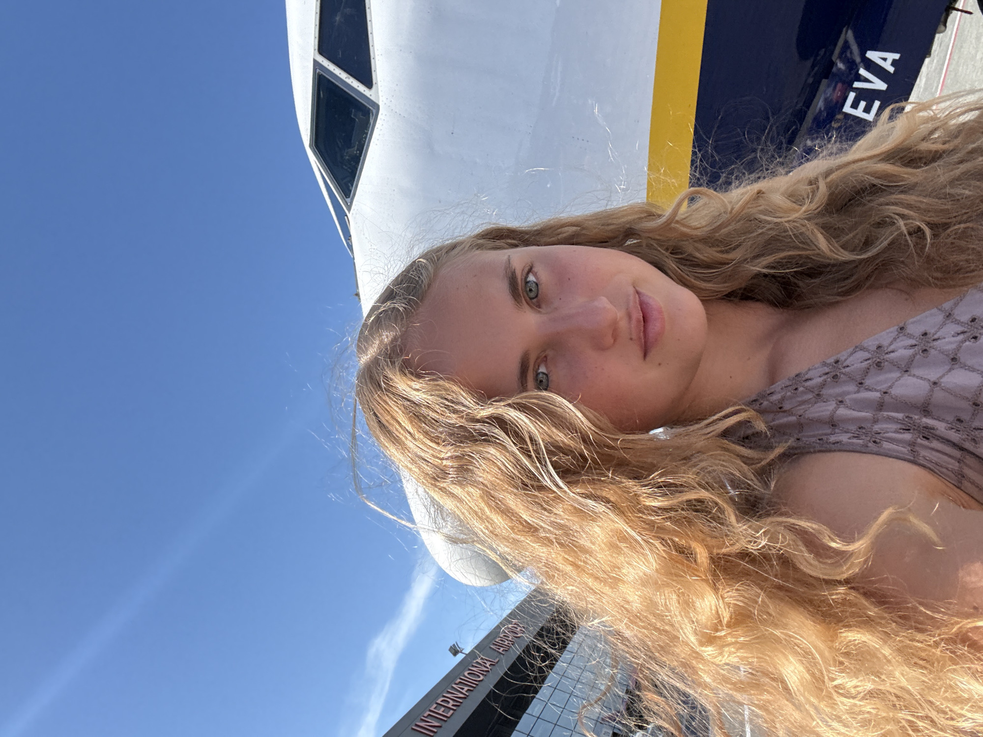 A young Caucasian woman with long, curly blonde hair and blue-green eyes is taking a selfie outdoors. She is wearing a light purple sleeveless top with a patterned texture. Behind her, a large white airplane with a dark blue fuselage and a yellow stripe is partially visible. The plane's cockpit windows are prominent. To the left, the exterior of a modern building with glass windows is visible, featuring red lettering that reads "INTERNATIONAL AIRPORT". The sky above is a clear, bright blue. The scene appears to be at an airport.
