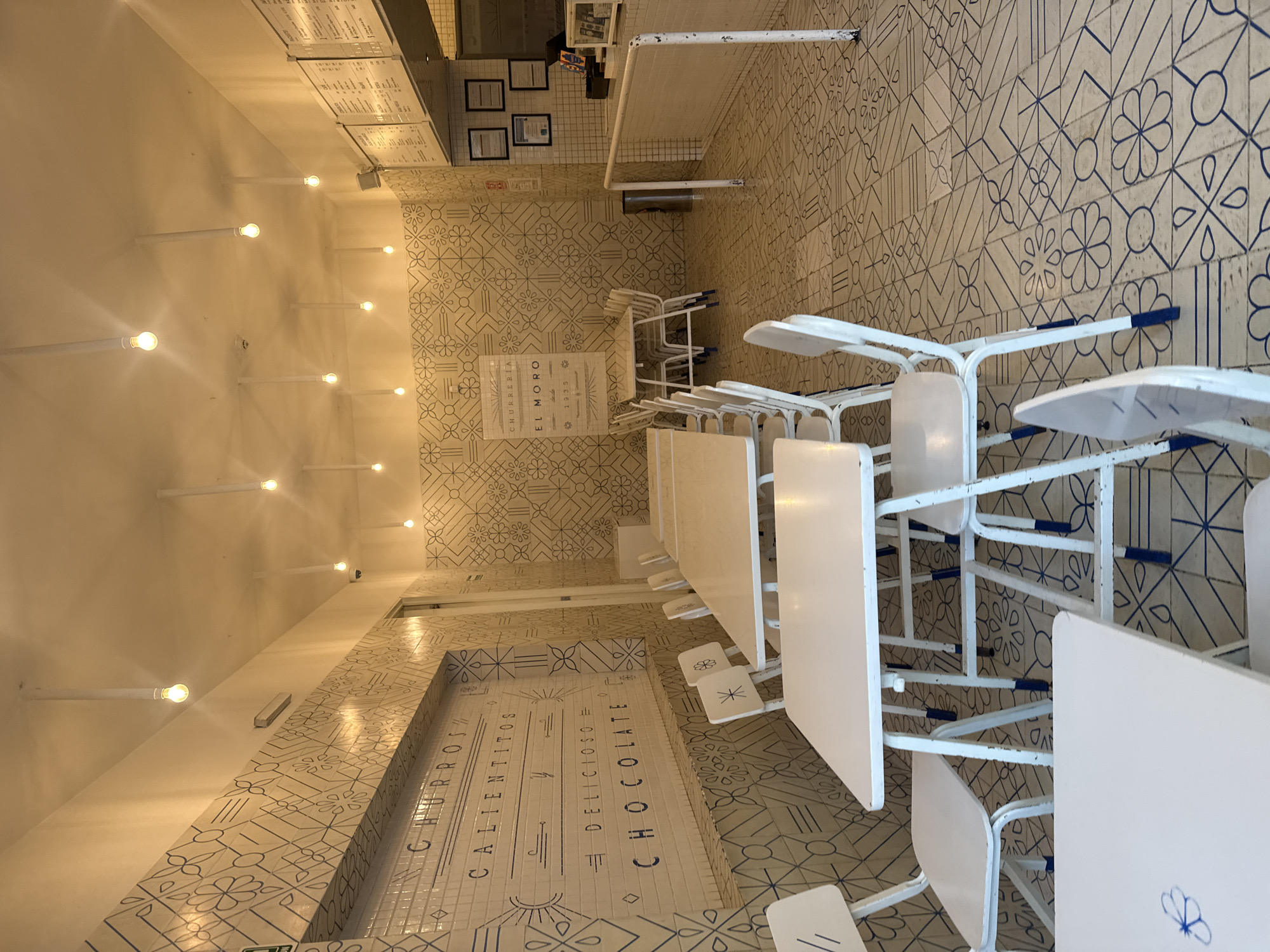 The image shows the interior of a churrería, most likely El Moro in Mexico City, Mexico. The space features white walls and floors with blue geometric patterns. A series of long, narrow white tables with matching chairs are arranged in rows. The chairs are white with blue accents. The walls are decorated with lettering and designs reminiscent of tilework. There are several evenly spaced lights extending from one wall to the other. The space appears empty of customers, and the scene is brightly lit. A menu board is visible near the serving area.