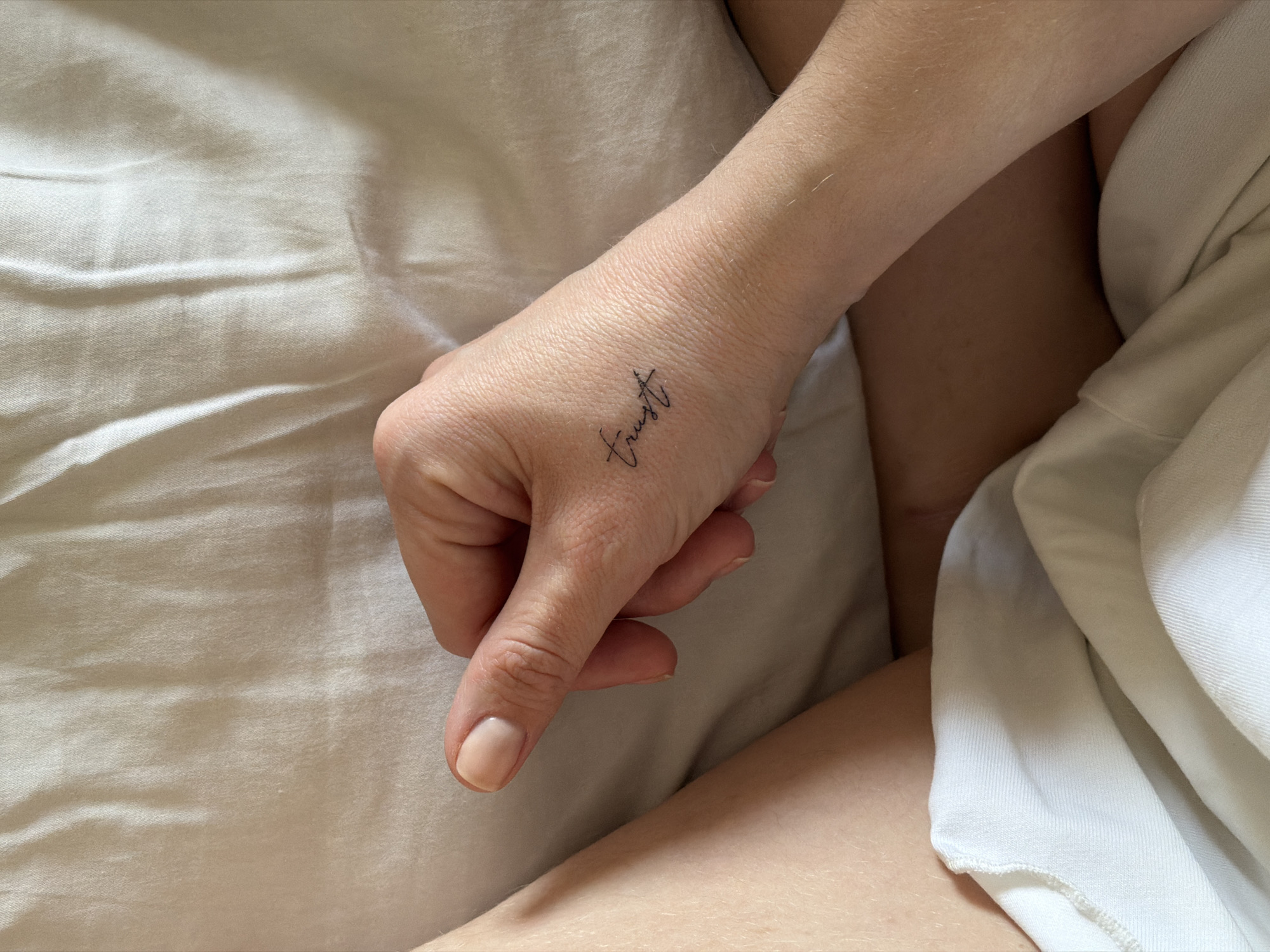 The image captures a close-up, overhead perspective of an individual's right hand and lower body, set against a backdrop of wrinkled, light-colored fabric consistent with bedding. The hand rests palm down, exhibiting a natural nail color with a light nude or clear polish. A small, black script tattoo, spelling "trust," is present on the dorsal surface of the hand near the wrist. Portions of the person's upper thighs are visible, partially obscured by a section of white fabric, likely part of clothing or additional bedding. The scene is observed indoors, in a residential or private setting, located in Khimki, Russia.