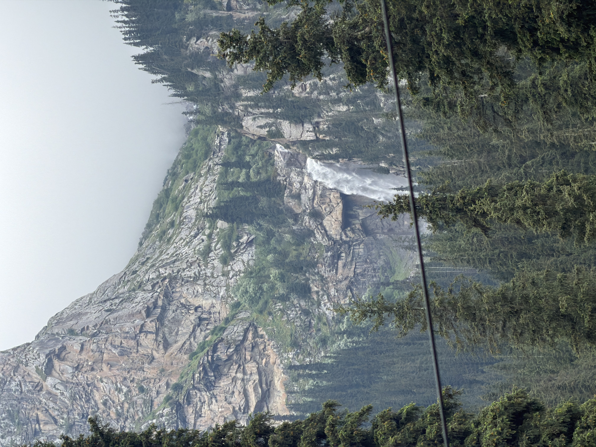 A waterfall cascades down a rocky mountainside surrounded by a dense forest of evergreen trees. The sky is overcast, and the overall atmosphere is damp and misty. The image captures a natural landscape, likely in a mountainous region of Kyrgyzstan, given the reference to an unknown city within that country. The foreground shows the tops of several tall evergreen trees and a dark horizontal line, possibly a power cable, stretching across the frame. The middle ground is dominated by the waterfall and the rugged, textured surface of the mountain, interspersed with patches of green vegetation and more trees. The background is a hazy expanse of mountains and sky, obscured by mist and clouds. There are no people or animals visible in the image.