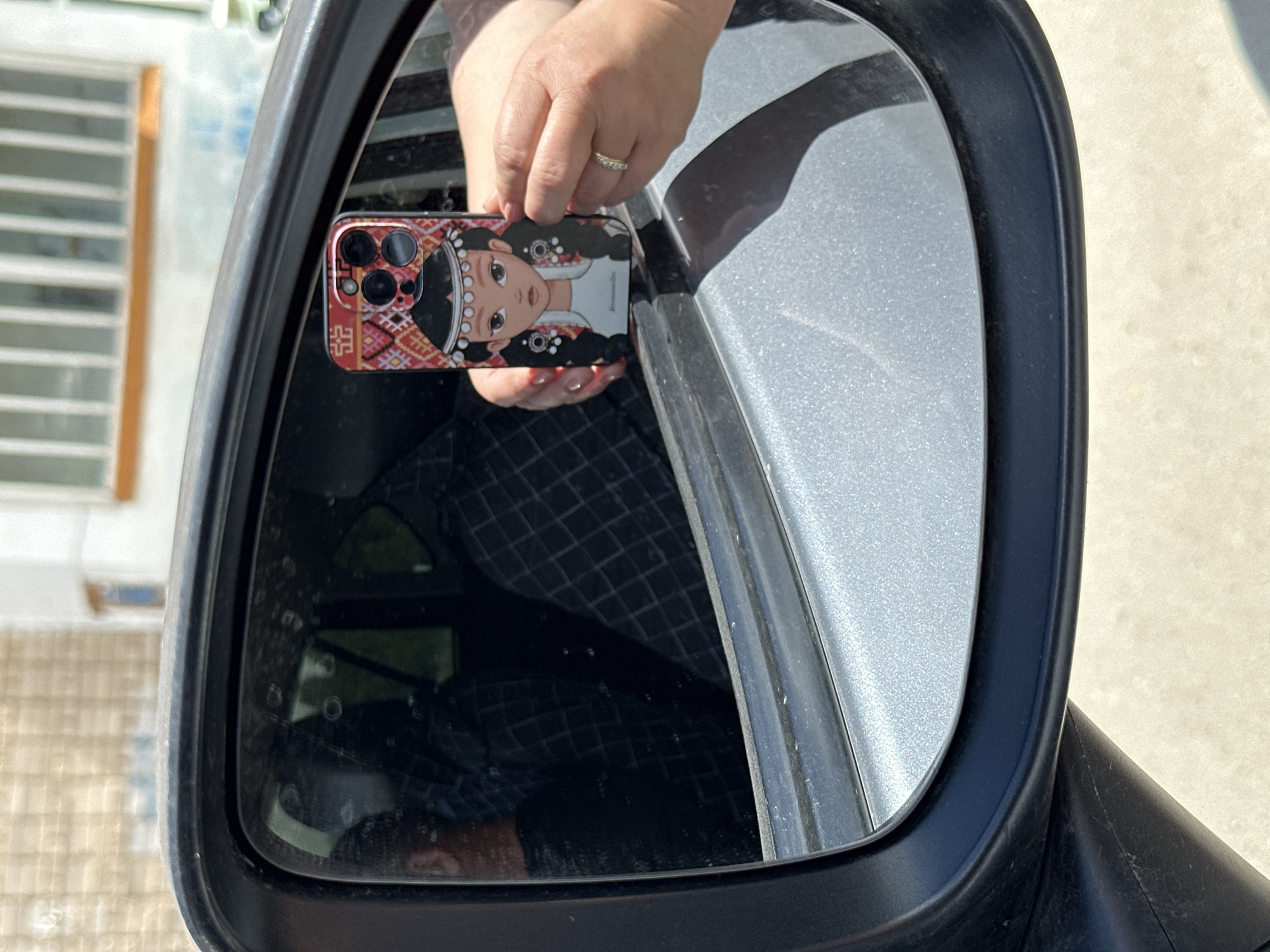The image shows a reflection in a car's side mirror. In the reflection, a person is holding up a smartphone with a decorative case featuring an illustration of a young woman with traditional adornments. The phone case has a red and white patterned border and a depiction of a woman with dark hair, adorned with pearls and earrings. The person holding the phone is wearing a ring on their left hand. Inside the car, visible in the mirror, are two individuals, one in the driver's seat wearing a dark checkered shirt. The background of the reflection shows a building with barred windows. The car mirror is attached to the car's exterior, which is gray. The scene appears to be outdoors, with sunlight illuminating the area. The location is the city of Uchaly, Russia.
