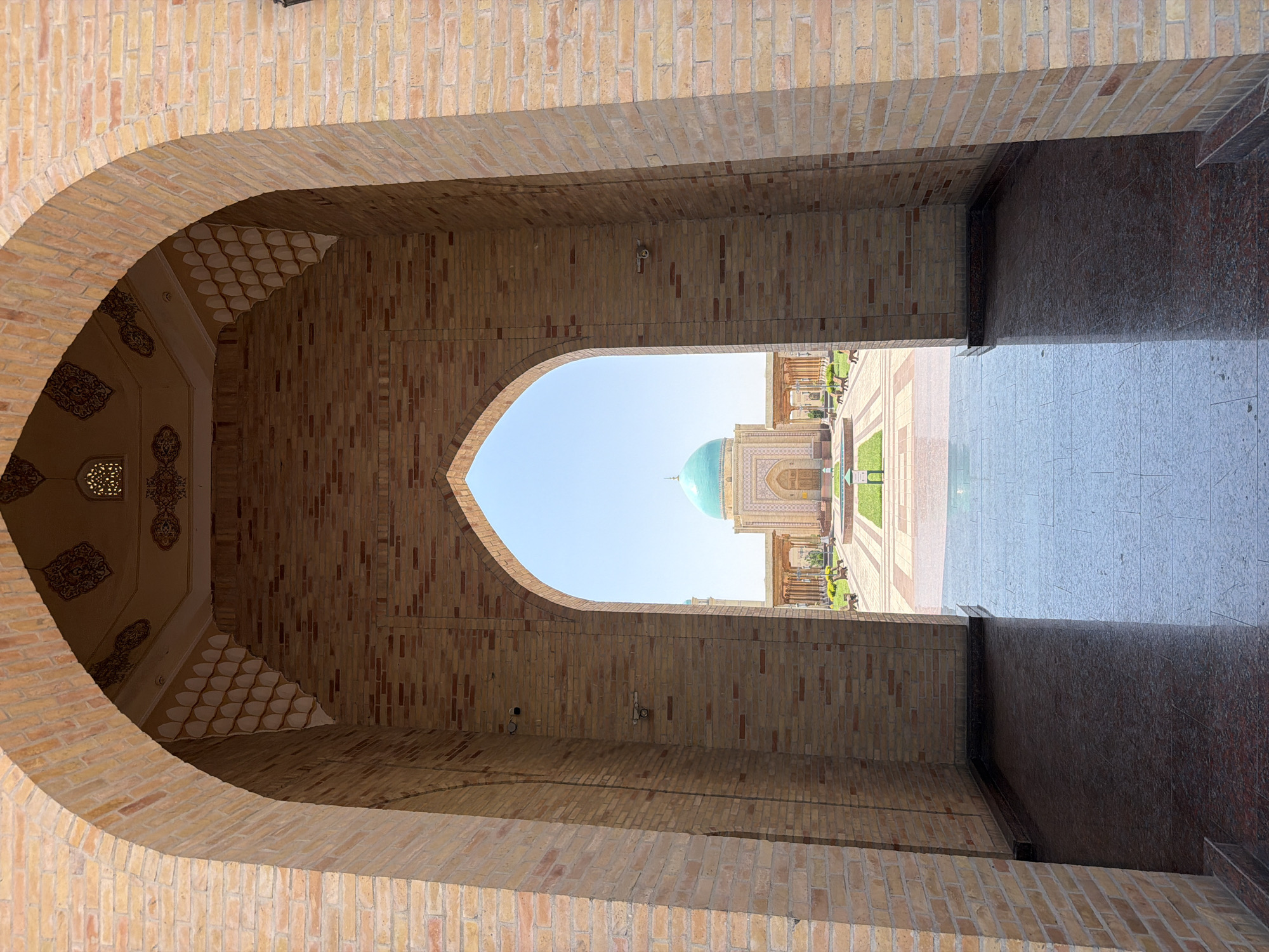 The image shows a view through a large, arched brick gateway towards a mosque with a turquoise dome under a clear blue sky. The gateway is constructed of light brown bricks and features decorative patterns on the interior ceiling. The ground inside the gateway is covered with dark, reflective flooring, likely granite.
Beyond the gateway, a courtyard unfolds. This courtyard is paved with patterned tiles and features manicured green lawns, benches, and trees. In the center of the courtyard, a path leads towards the mosque. The mosque itself is a light-colored building with intricate architectural details and a prominent turquoise dome.
The location is the Tashkent Islamic University in Tashkent, Uzbekistan. This institution is known for its impressive architecture, which blends traditional Islamic design with modern elements. The university campus is a significant landmark in the city, reflecting the importance of Islamic scholarship and culture in Uzbekistan. The scene captures a serene and majestic atmosphere, typical of religious and academic institutions.