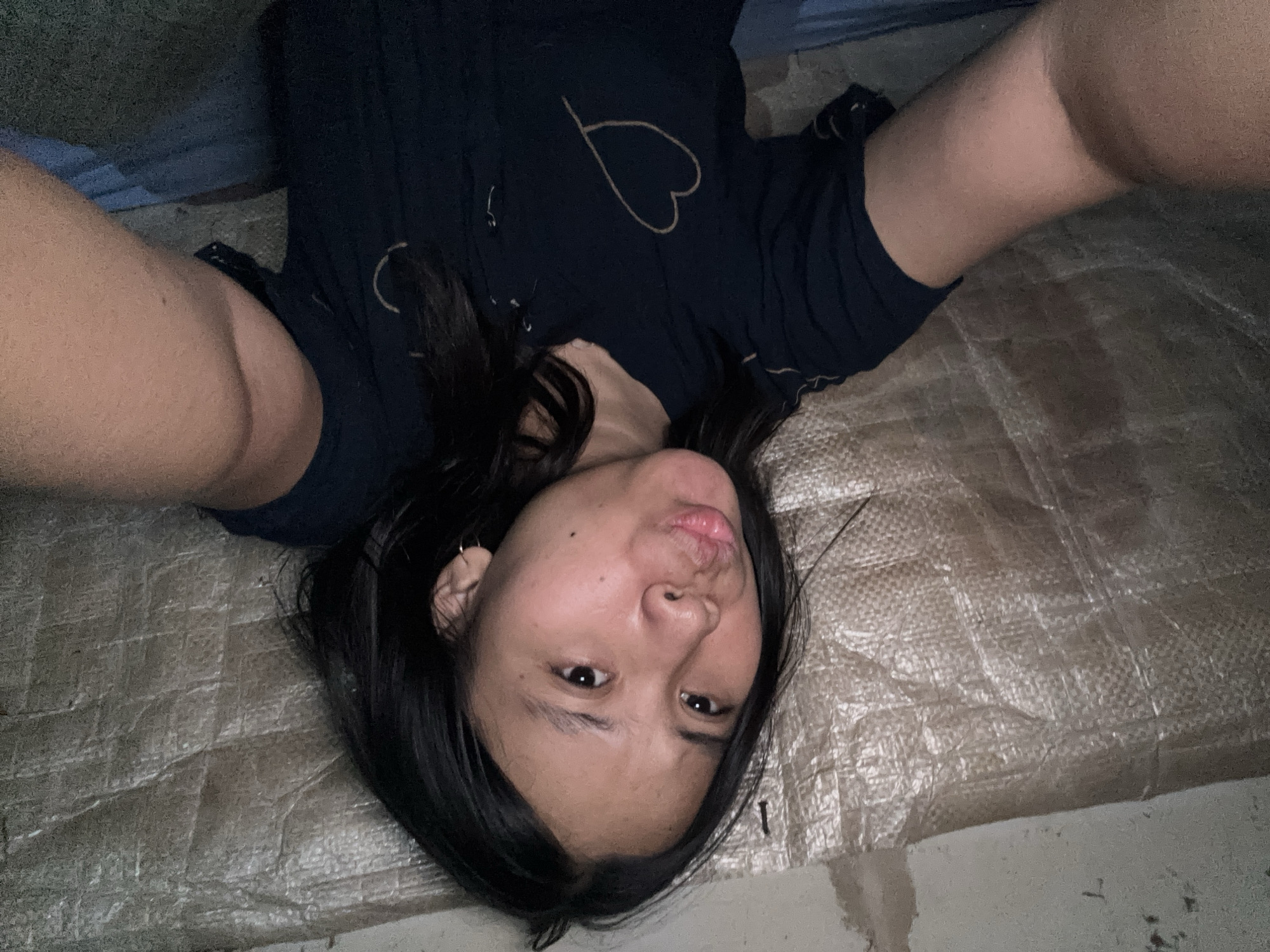 A young woman with dark hair and light skin is taking a selfie. She is wearing a dark blue t-shirt with a heart design on it. She is making a kissy face. The background appears to be a textured wall or tarp. The lighting is dim and casts shadows. The scene is indoors. The location is the city of East Jakarta, Indonesia.