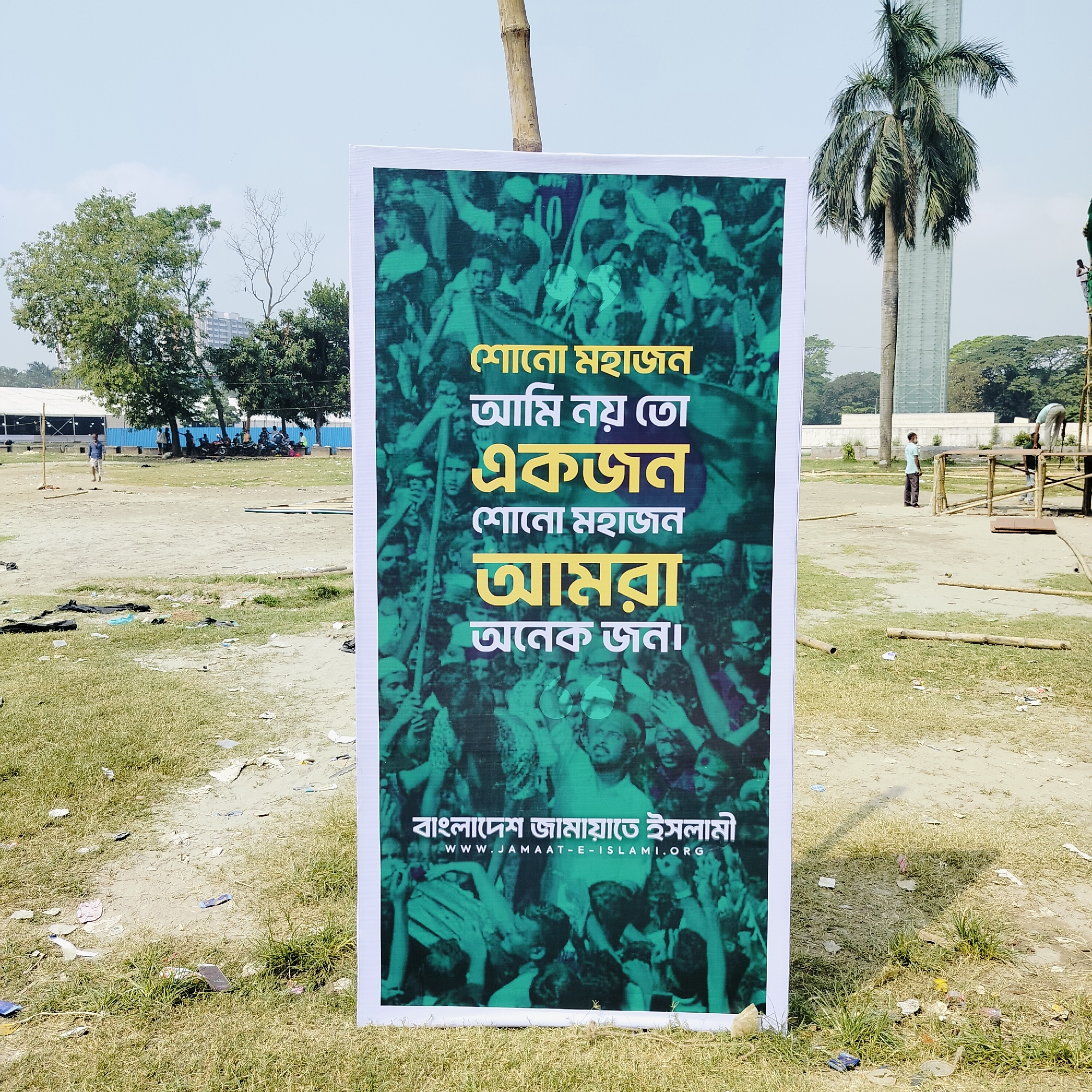 The image shows a poster for Bangladesh Jamaat-e-Islami, an Islamist political party in Bangladesh. The poster is displayed in a field, likely in Dhaka, Bangladesh. The poster's text includes the party's name and website address. The background of the poster features a green-toned image of a crowd. In the background of the image, there are people, trees, and a partially constructed building.