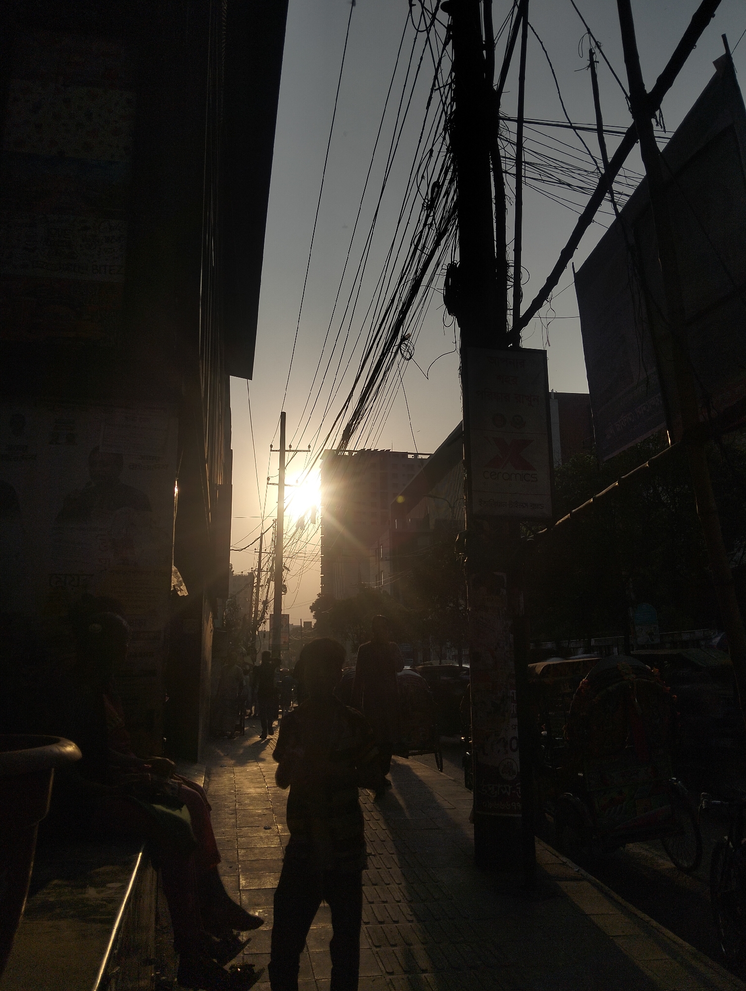 The image depicts a street scene in Dhaka, Bangladesh, during sunset. A mix of pedestrians and cycle rickshaws are present on the right side of the frame. A person is standing in the foreground. Silhouetted figures are sitting on a ledge on the left. Above, a dense network of electrical wires and poles obscures the sky. Advertisements are visible on building facades. The setting sun is between two buildings in the distance.