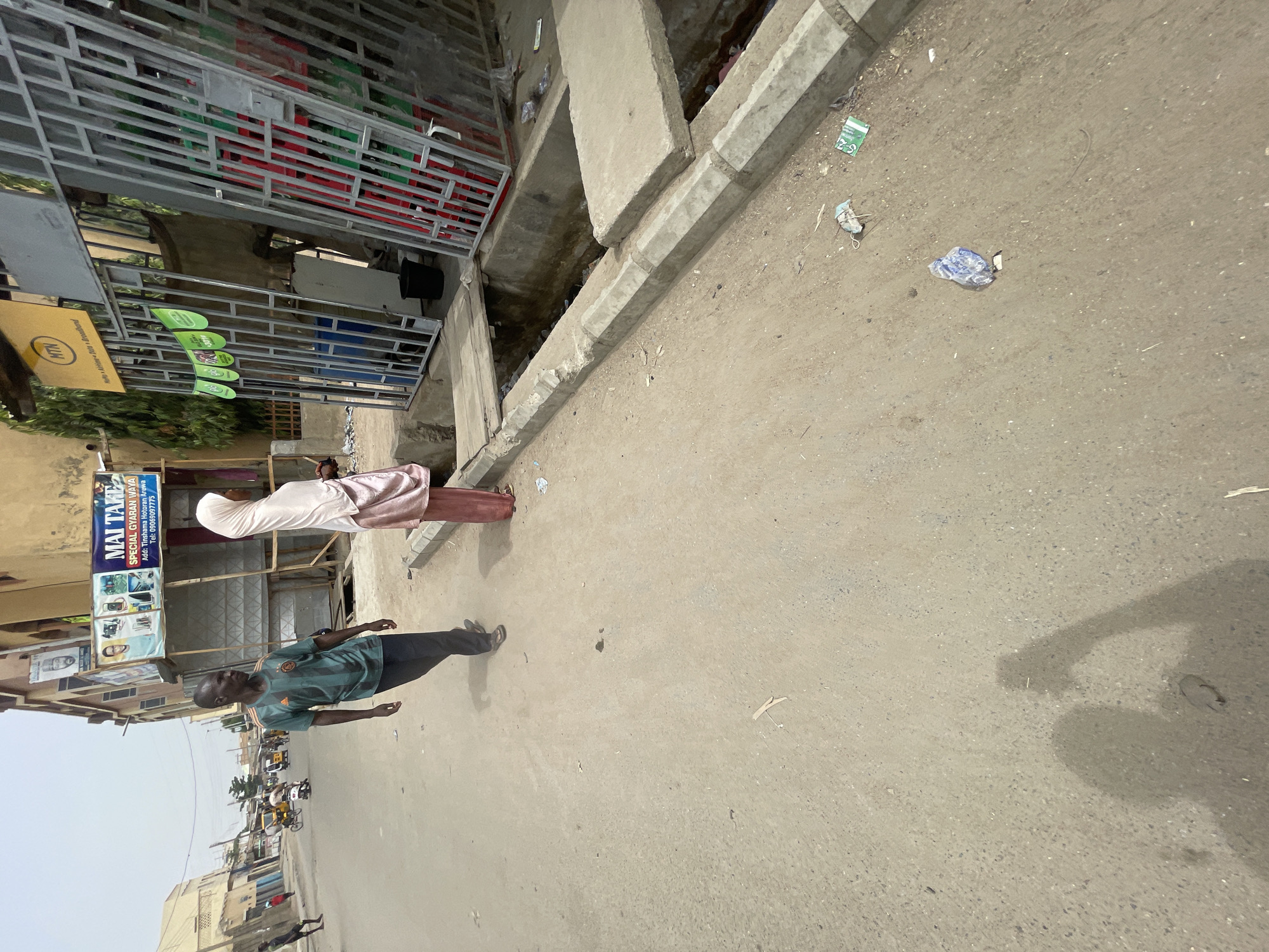 The image depicts a street scene in an urban area. A man in a green shirt and dark pants is walking on the street. Next to him, there's a woman wearing a light-colored dress with a head covering. The setting appears to be a commercial district with buildings, shops, and signage. There's an advertisement reading "MAI TAKE SPECIAL GYARAN WAYA" and a phone number, which indicates a business specializing in phone repairs. A sign with "MTN" is also visible. The ground is littered with some debris. Traffic is seen in the distance, suggesting a populated area. The sky is overcast, providing soft lighting to the scene.