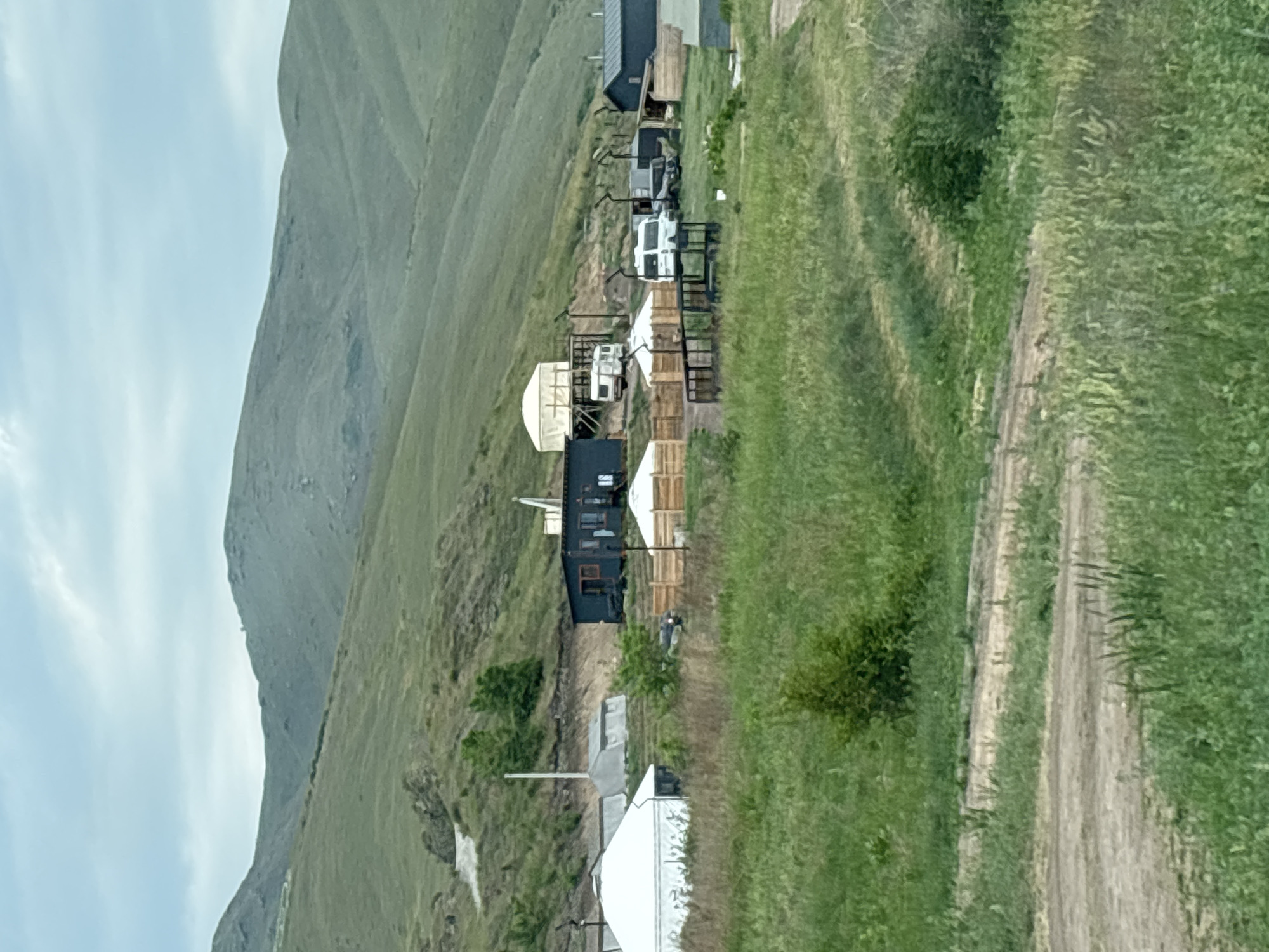 The image depicts a rural landscape with a cluster of buildings and tents situated on a grassy hillside. In the foreground, dirt paths wind through tall green grass. In the mid-ground, there are several structures including white tents, a dark-colored building with windows and a door, and a larger white yurt-like structure on a raised platform. Several vehicles, including a white camper van and what appears to be a darker SUV, are parked near the buildings. A wooden fence encloses a portion of the area. In the background, rolling green hills rise towards a hazy, overcast sky. The scene suggests a tourist or camping site in a natural, mountainous region. This location is identified as being in the city of Ust-Abakan, Russia, a region known for its natural beauty and mountainous terrain. There are no people visible in the image.