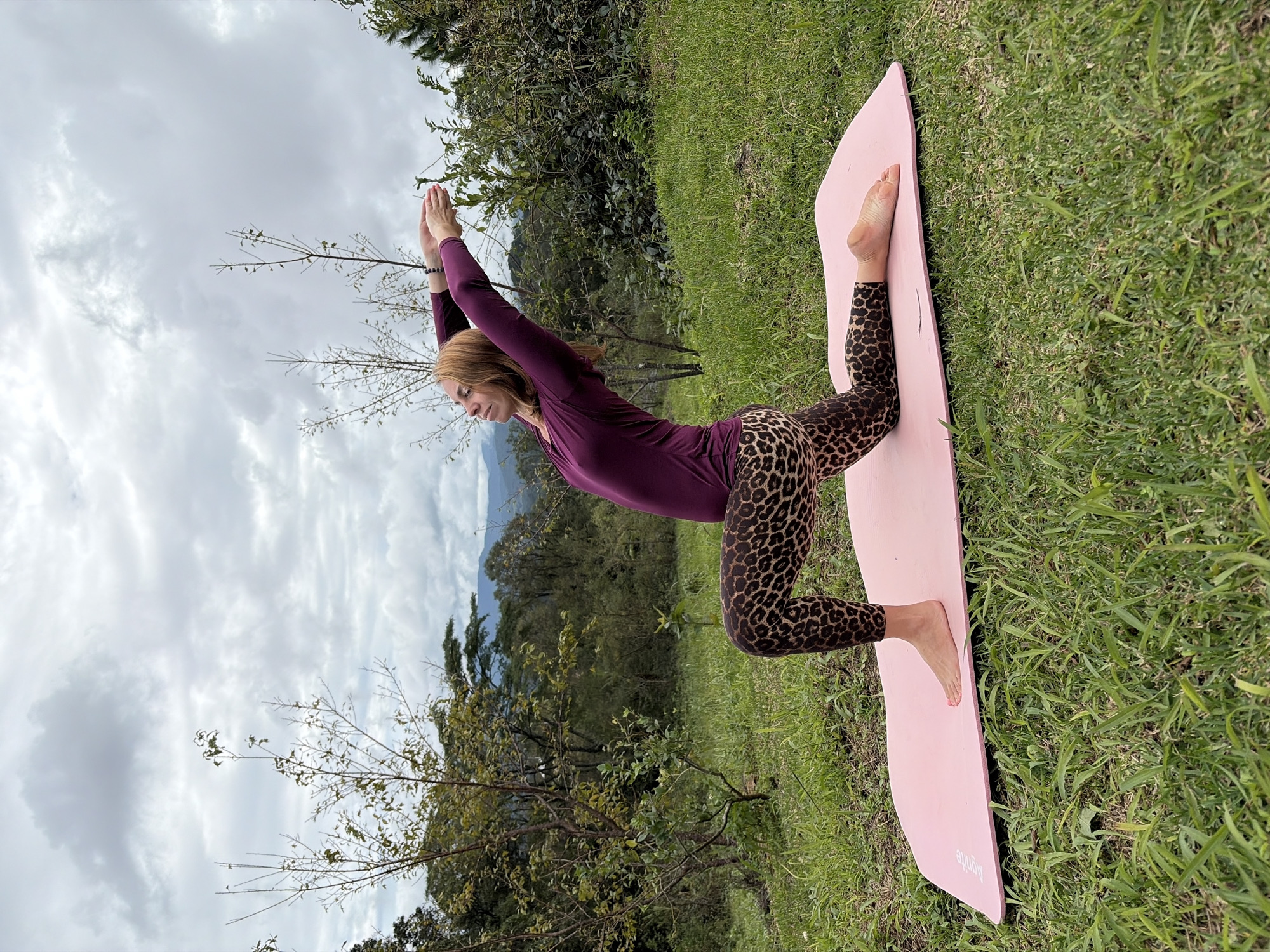 A woman with auburn hair is performing a yoga pose on a pink mat outdoors. She is wearing a plum-colored long-sleeved top and leopard-print leggings. Her right knee is on the mat, and her left leg is bent at a 90-degree angle with her foot flat on the mat. Her arms are raised above her head, with her hands clasped. She is facing to the left of the frame. The background shows a lush green hillside with trees and mountains in the distance under a cloudy sky. The location appears to be a natural setting, possibly in or near Ban Khun Wang, Thailand, given the vegetation and mountainous landscape.